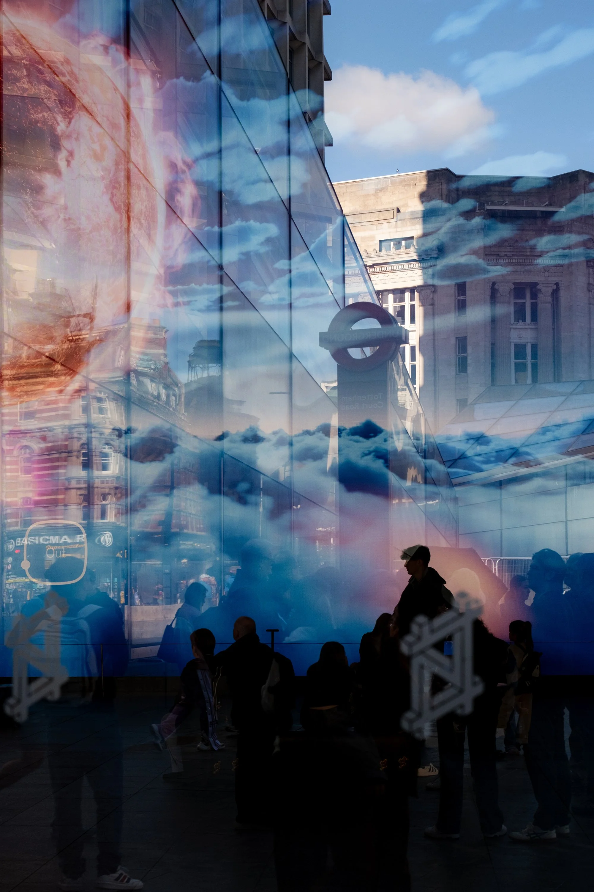 Reflections of clouds and city buildings on glass window, with silhouettes of people inside and outside.