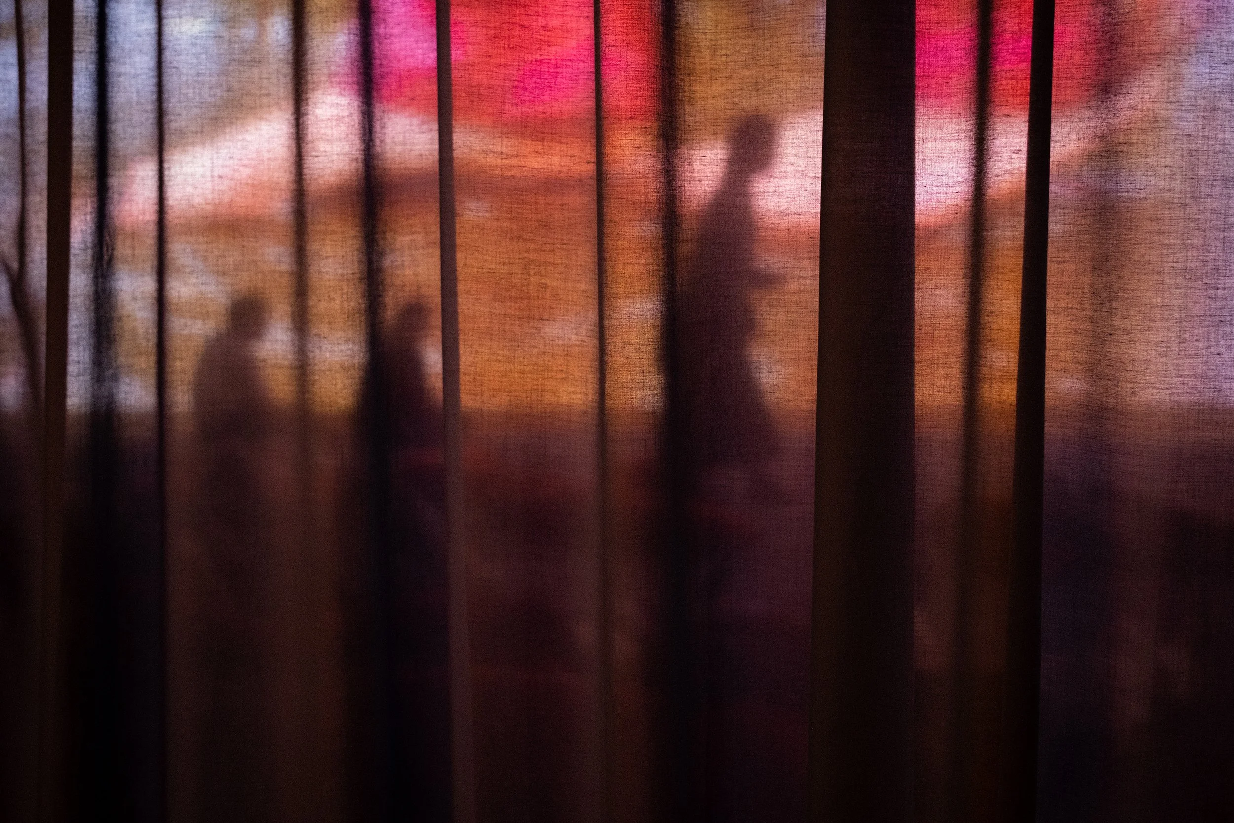 Silhouettes seen through a curtain, walking in front of a big screen showing bare feet of a person wearing red trousers. 