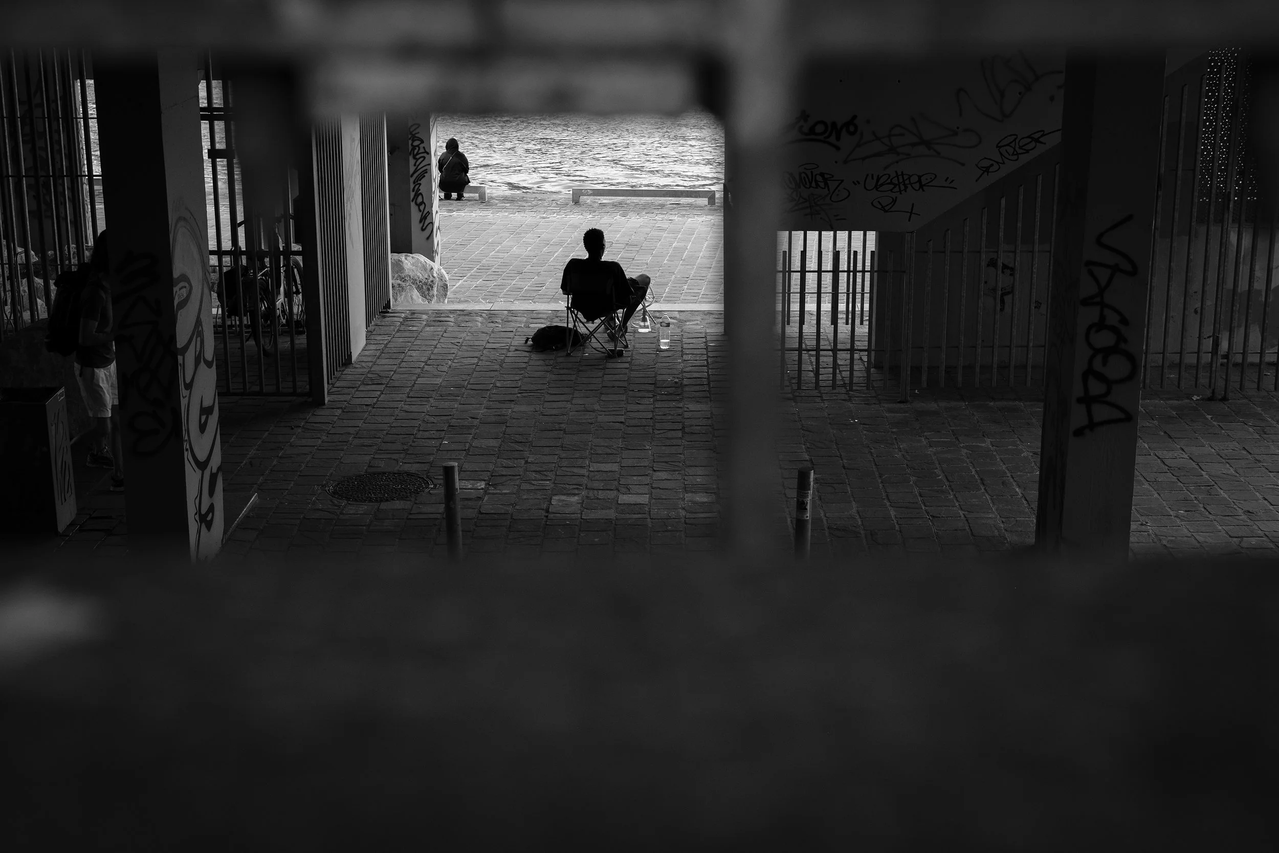 Black and white photo of a river scene viewed from under a bridge. Two people are present: one sitting in a camping chair near the water, another sitting at the bank of the river. The scene includes graffiti on the bridge supports and a cobblestone p
