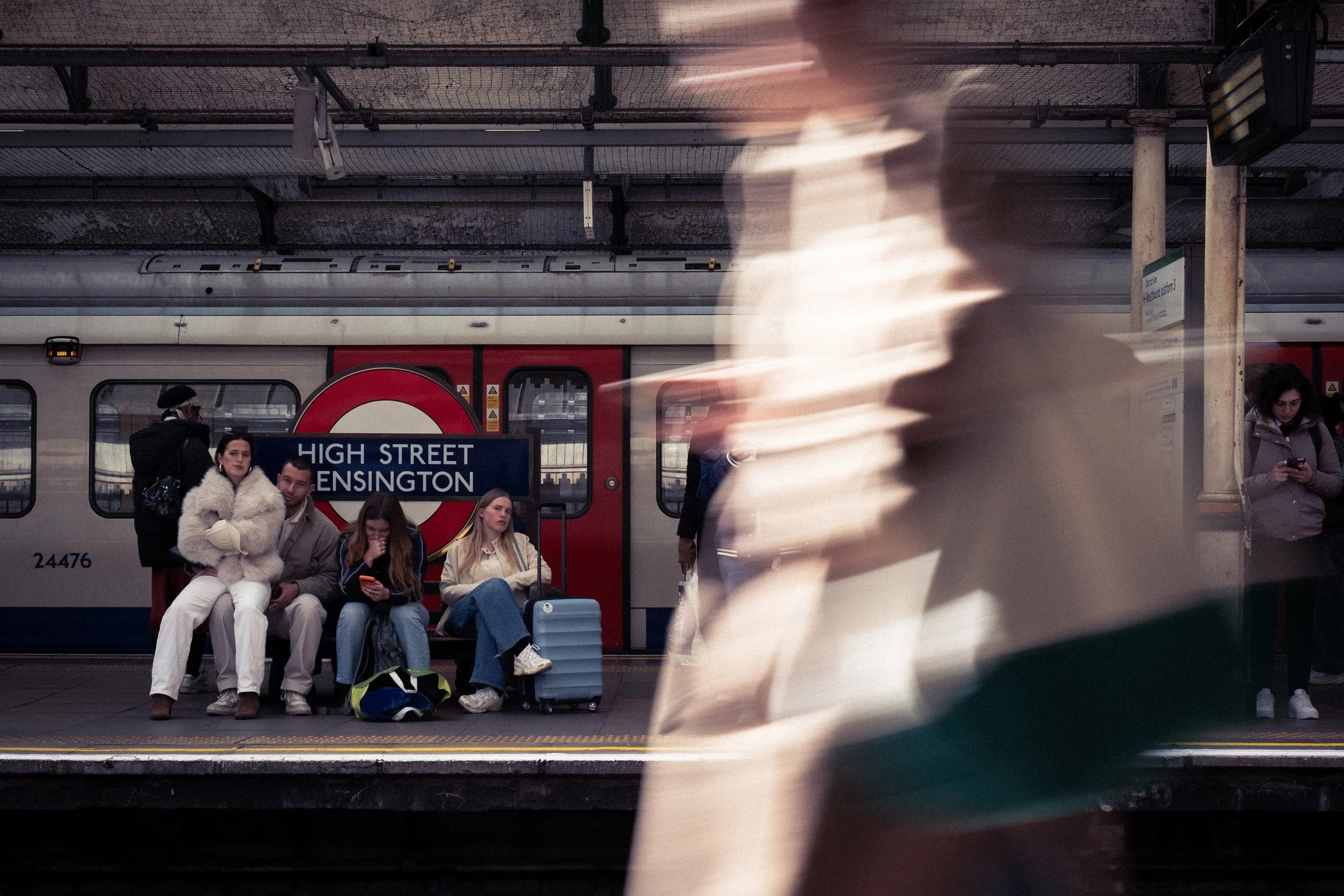 People waiting at a subway platform in front of a train, with a sign indicating it's the High Street Kensington station. Some are sitting, and some are using their phones. A blurred figure is walking past in the foreground.