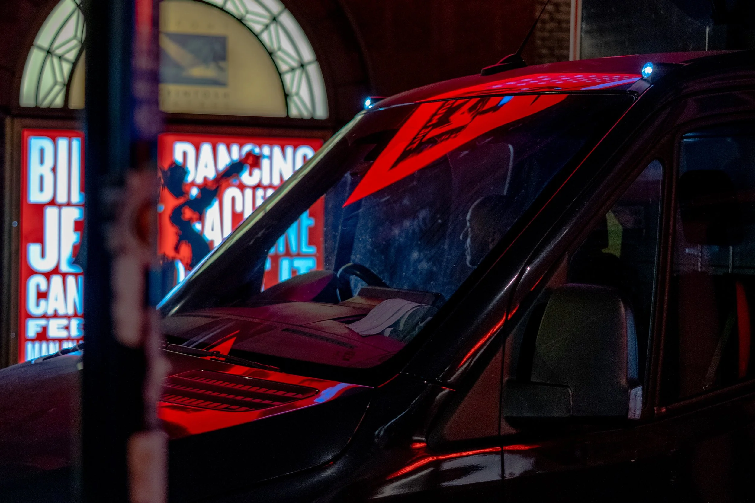 A black van with red graphical decals on the hood and roof, parked near neon-lit signs at night. The signs include a partially visible poster and a lit-up sign.