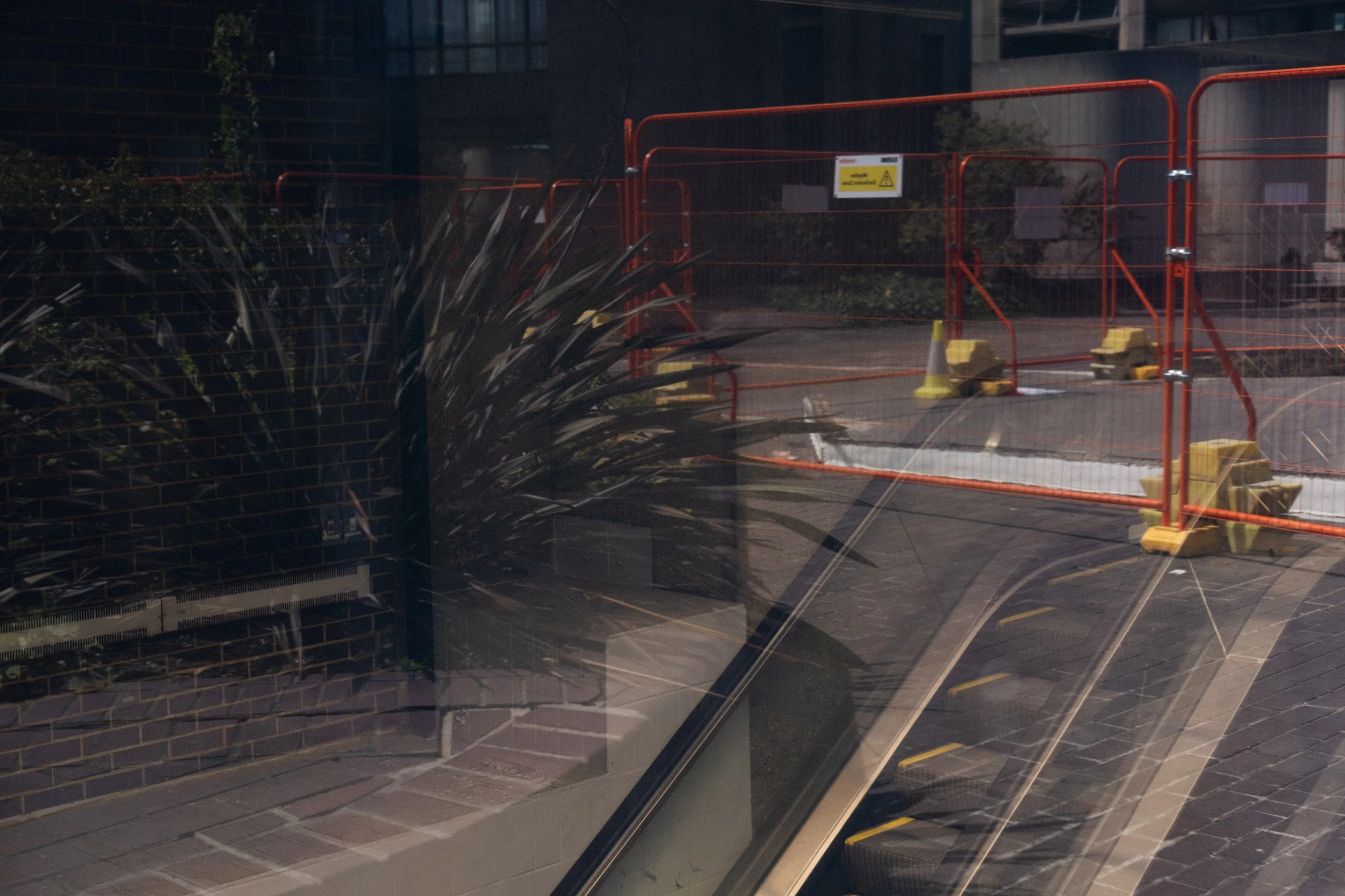 View of a construction site gated with orange fencing and caution signs, seen through a glass window with reflections of nearby plants and a brick building.