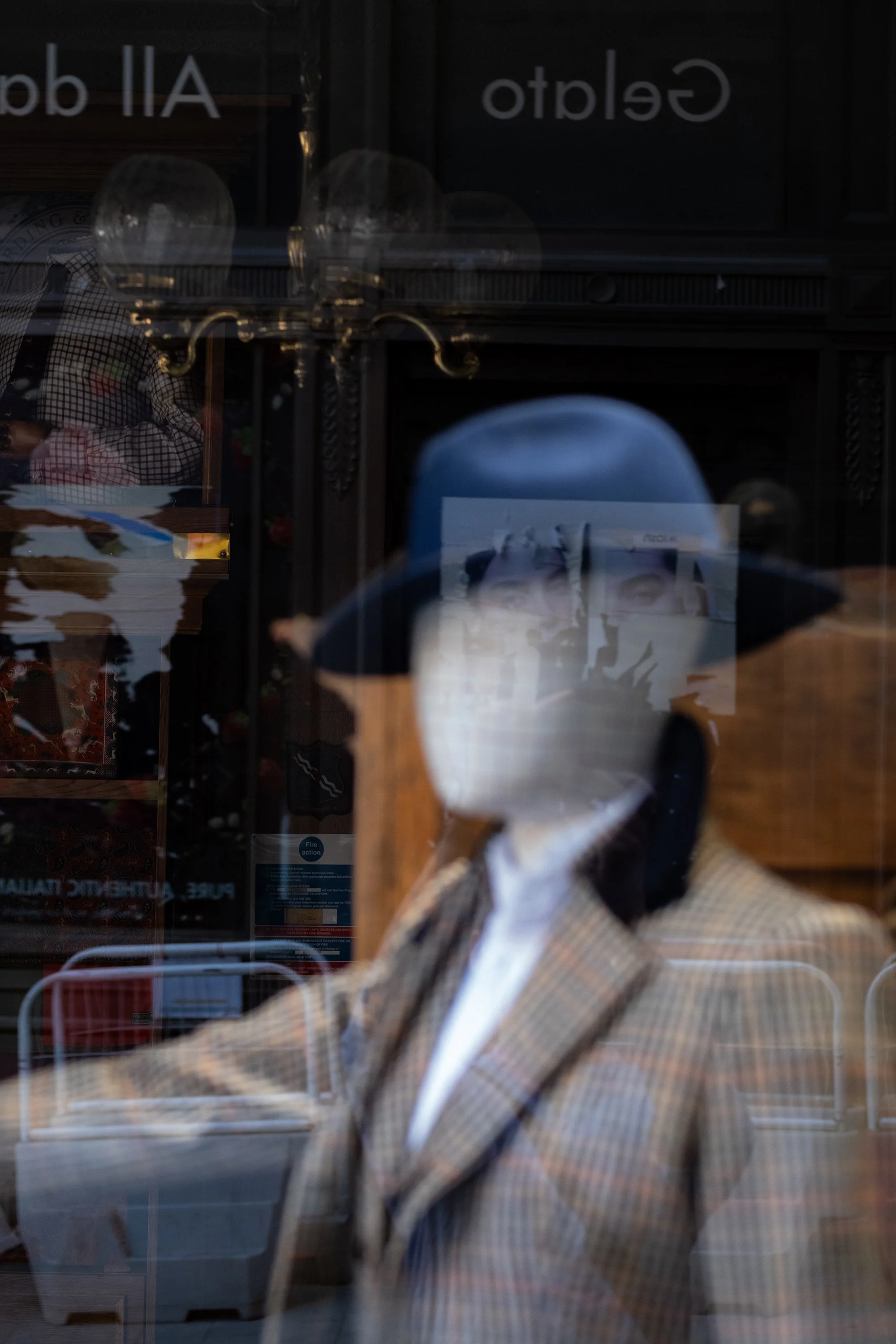 Reflection of a mannequin wearing a plaid blazer and a blue hat in a glass window, with indoor and outdoor elements visible.