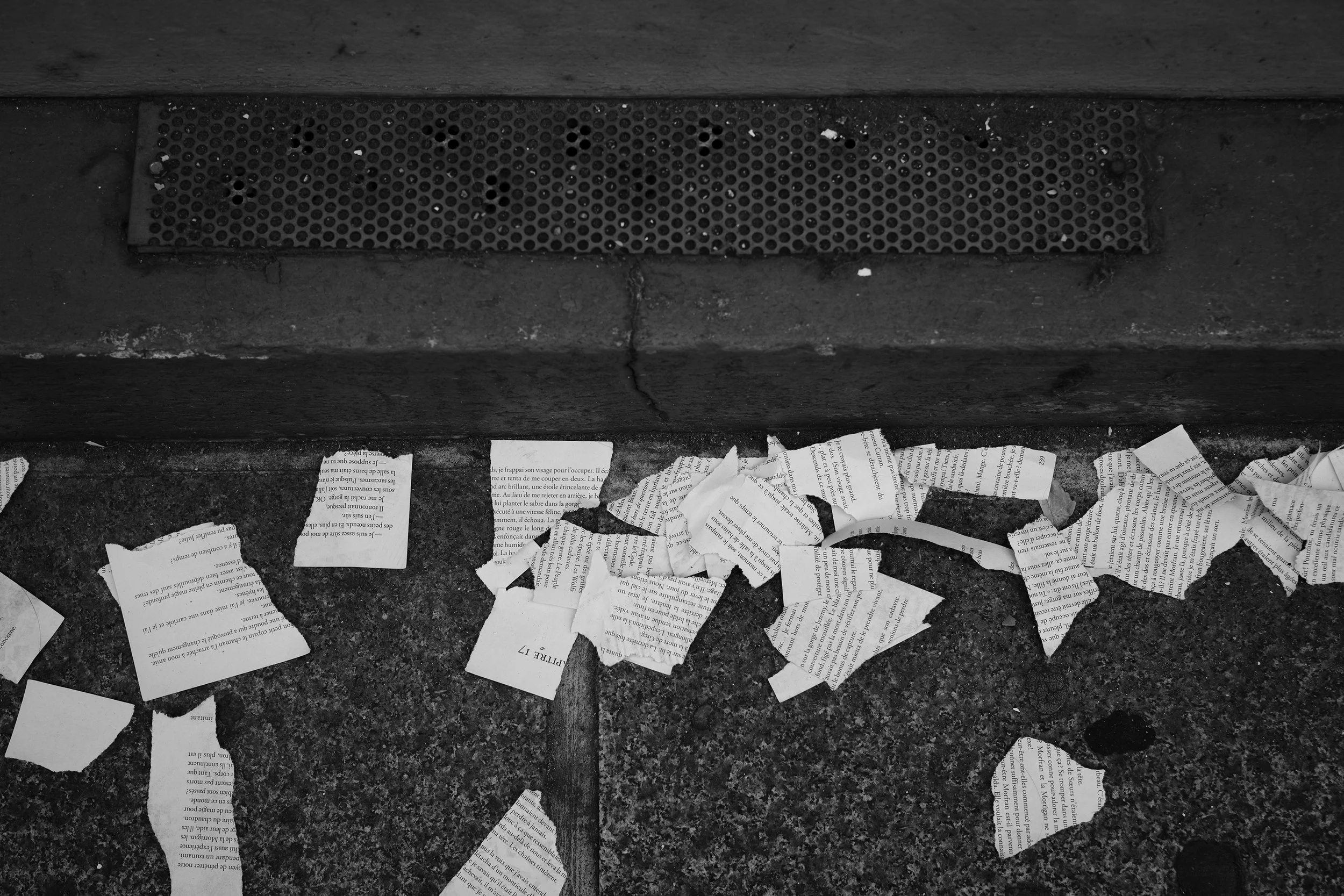 A black and white photo of torn pieces of paper scattered on a ground with a drain grate embedded in a concrete surface.