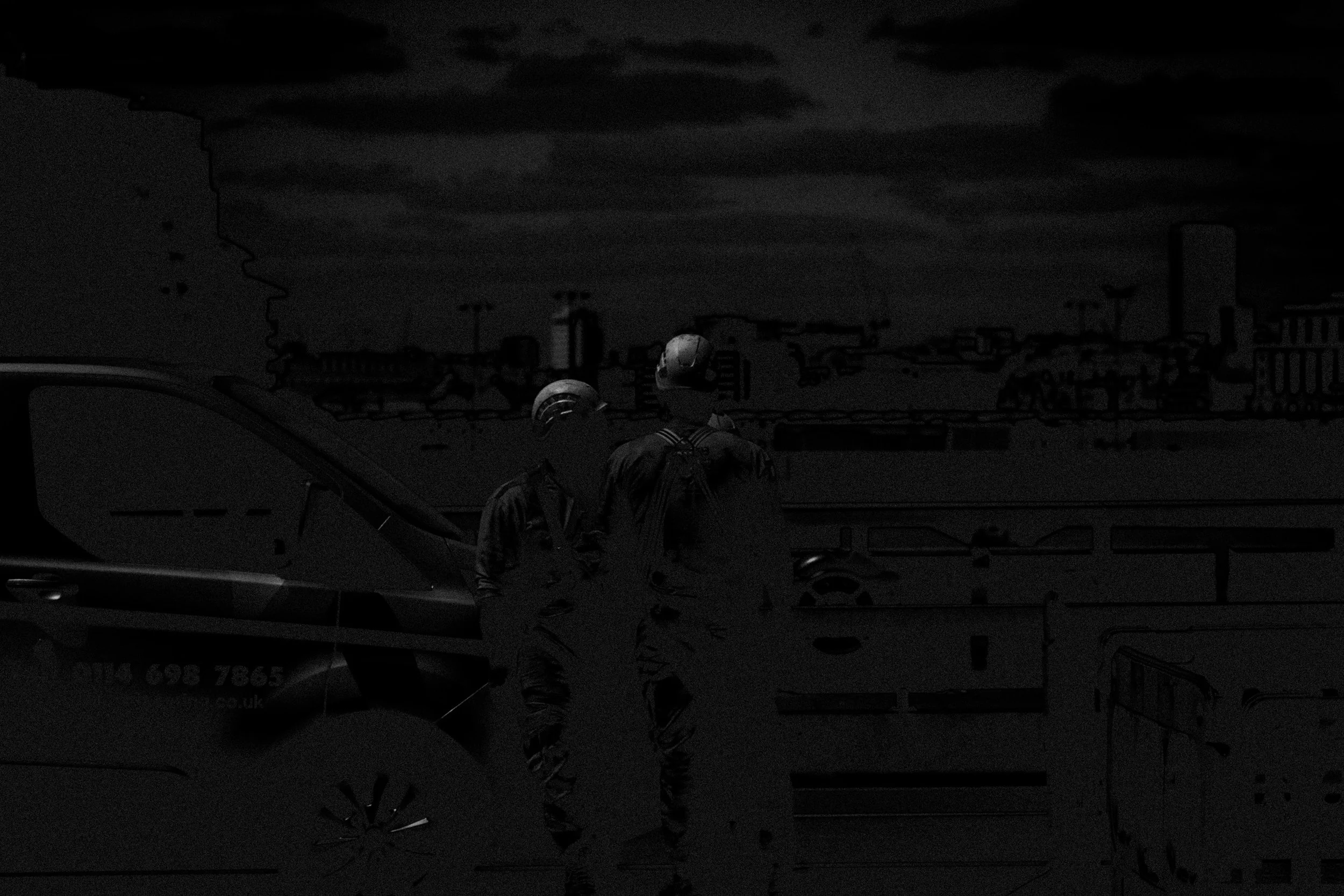 Dark abstract shot of two construction workers standing on a bridge, next to van, with the skyline of the City of London in the background.