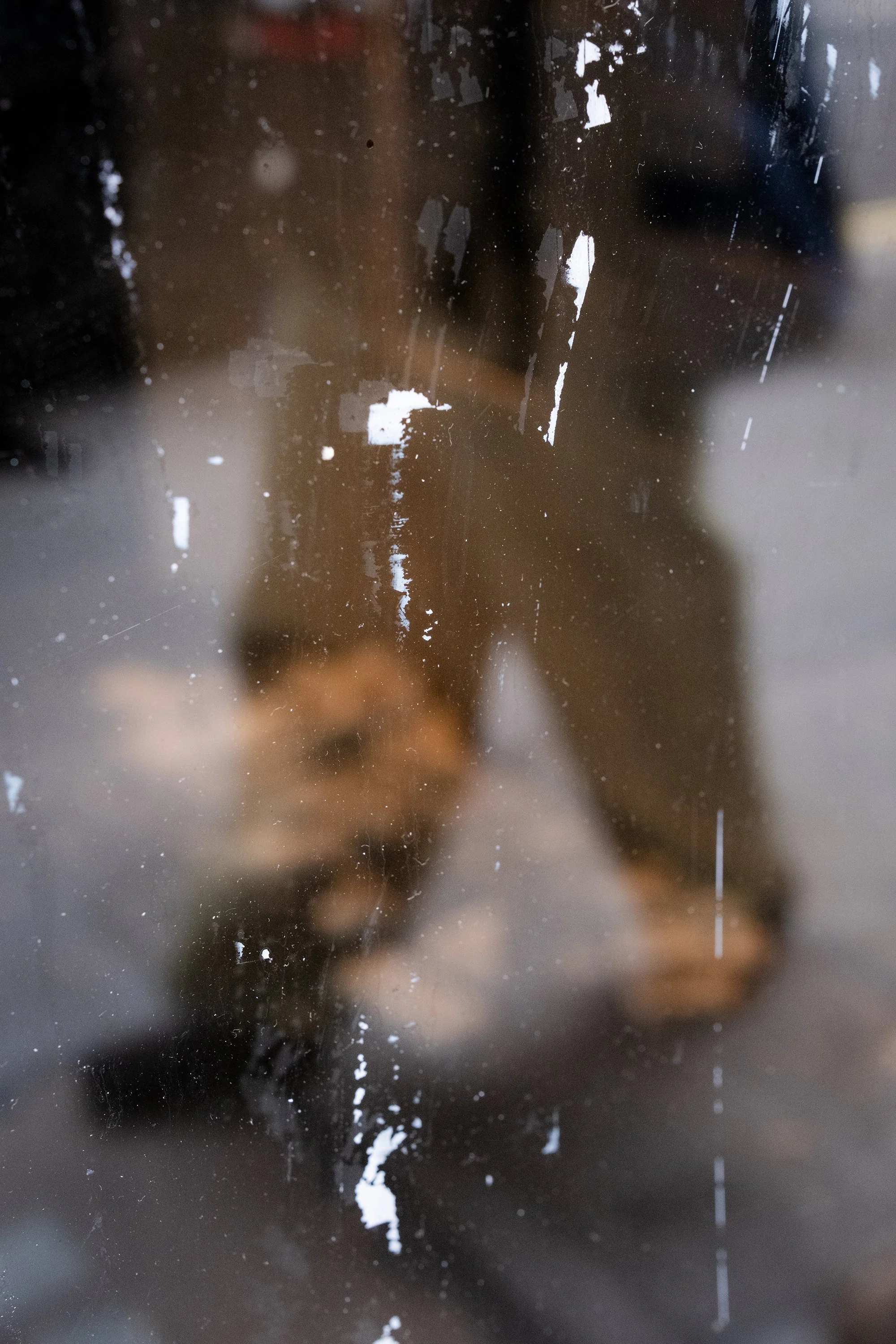 A dirty, scratched glass window with smudges and peeling paint, through which a person in dark clothing is taking a photo with a smartphone.