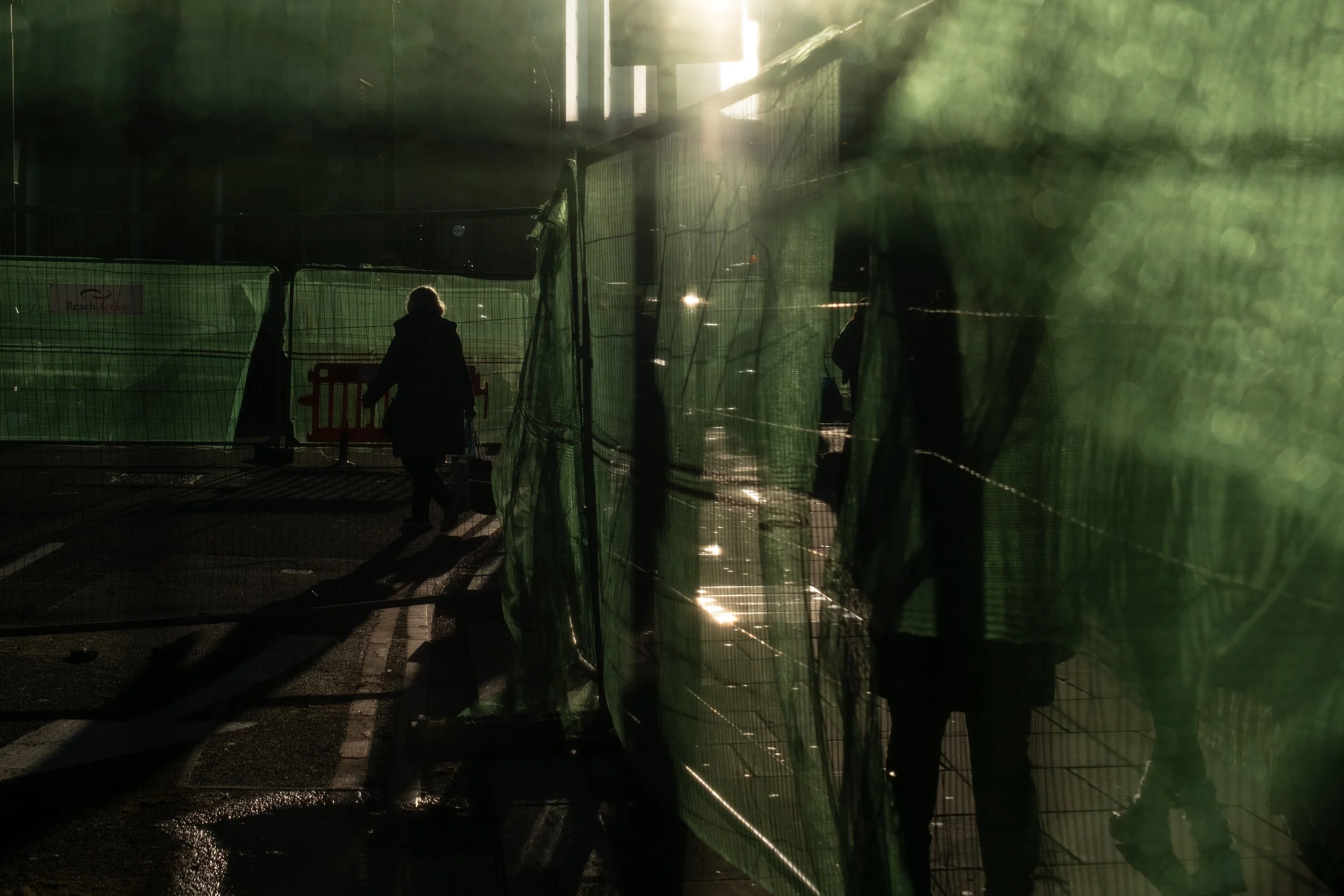 A person walking along a sidewalk with green construction fencing on the side, in sunlight creating lens flare and shadows.