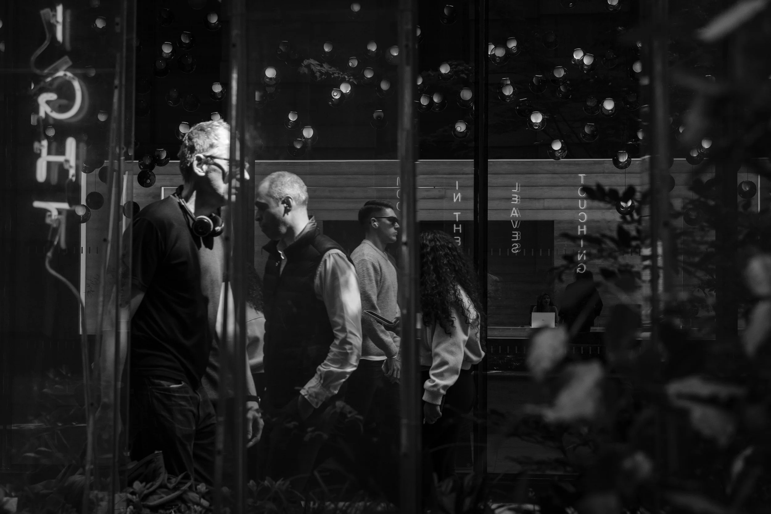 People walking outside, viewed through a glass window with reflections of lights and lettering, in a black and white photograph.