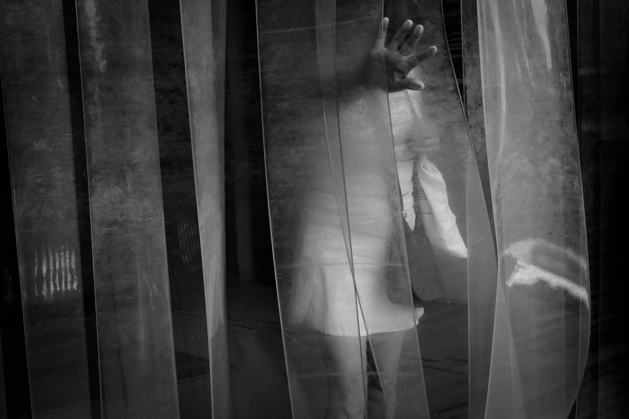 Black and white photo of a woman in a white dress walking through a PVC strip curtain, one hand pushing one of the strips out of her way.