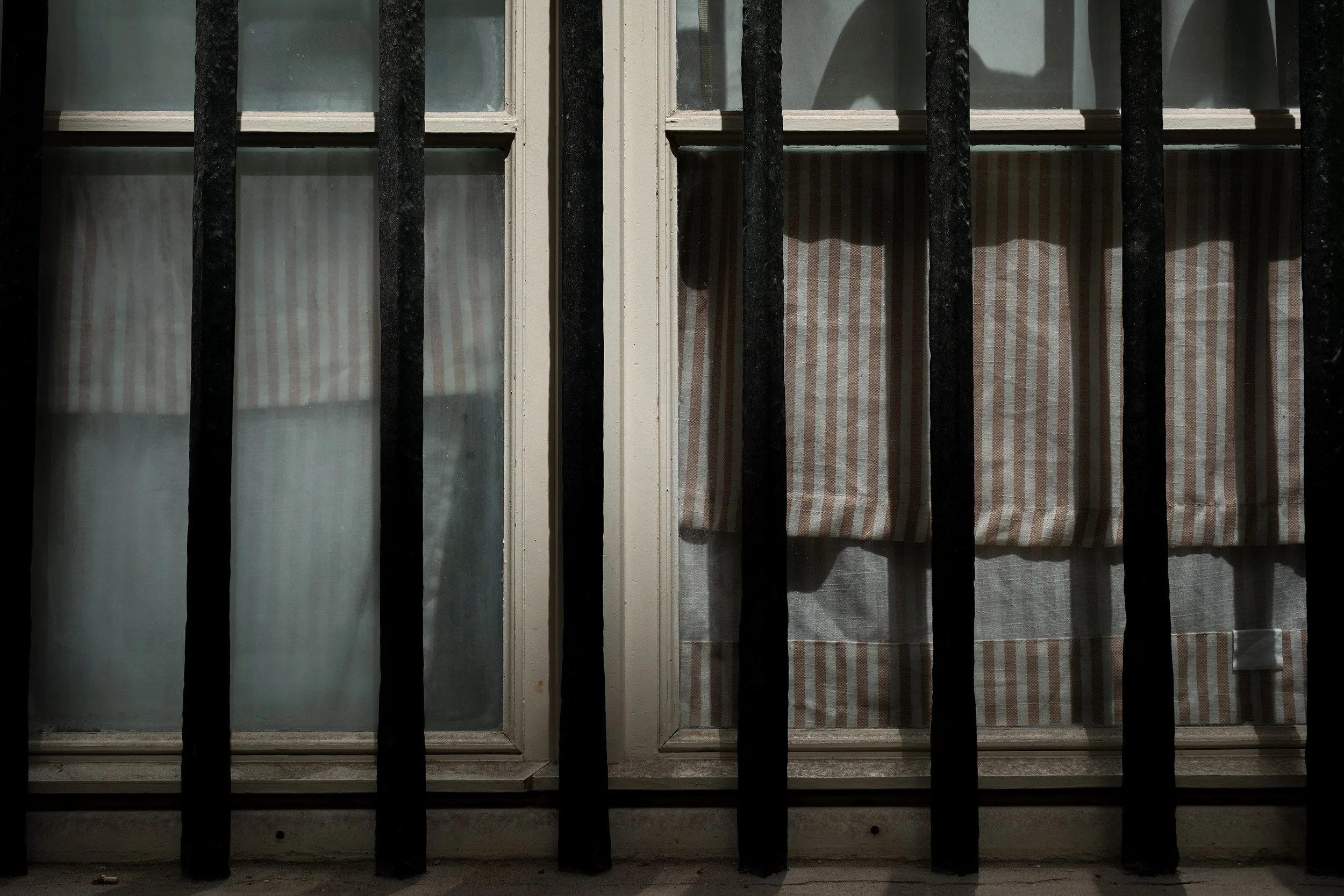 View of windows with striped curtains behind black metal bars, sunlight casting shadows on the curtains.