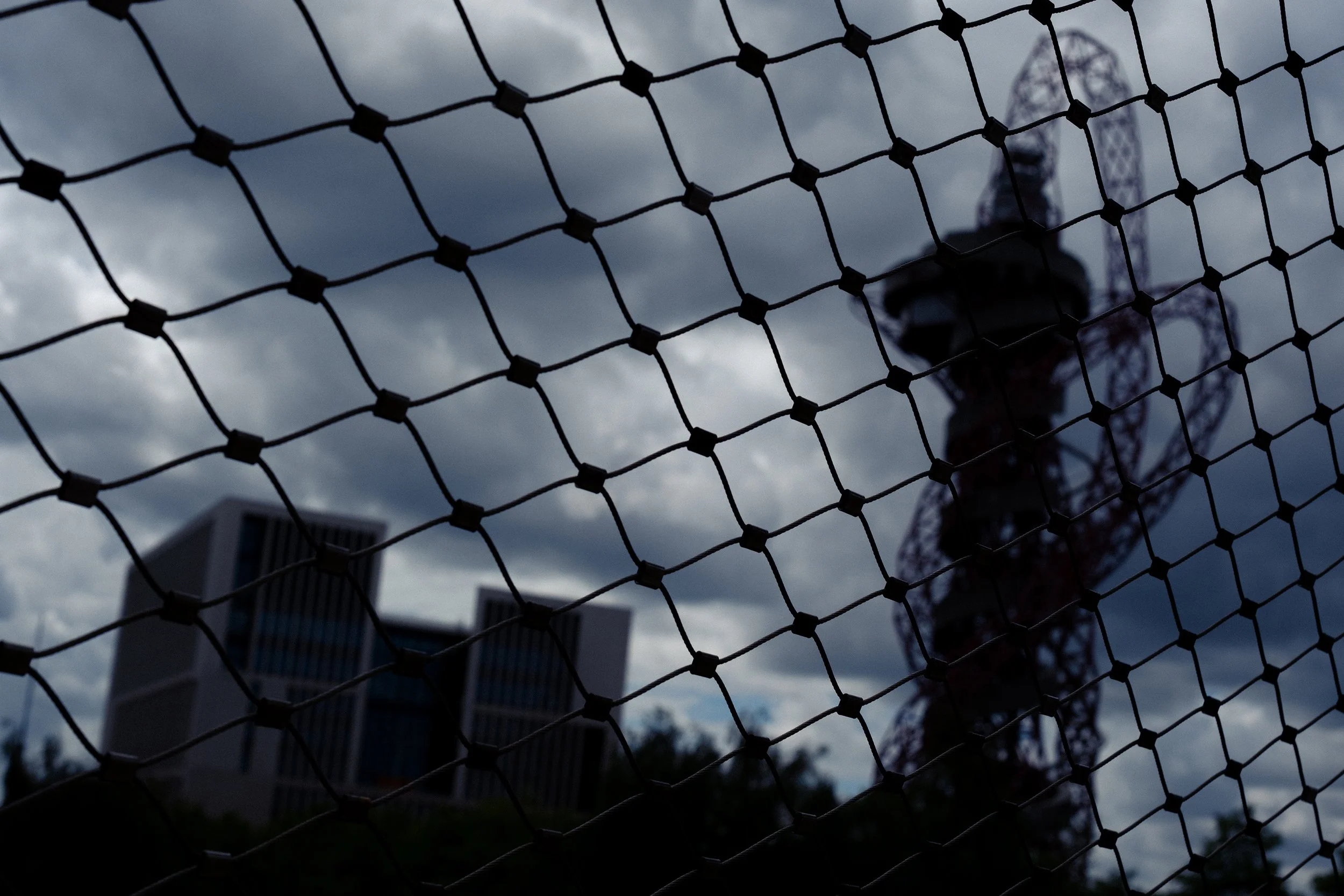 A view of the ArcelorMittal Orbit through a wire fence on a cloudy day with dark clouds in the sky.
