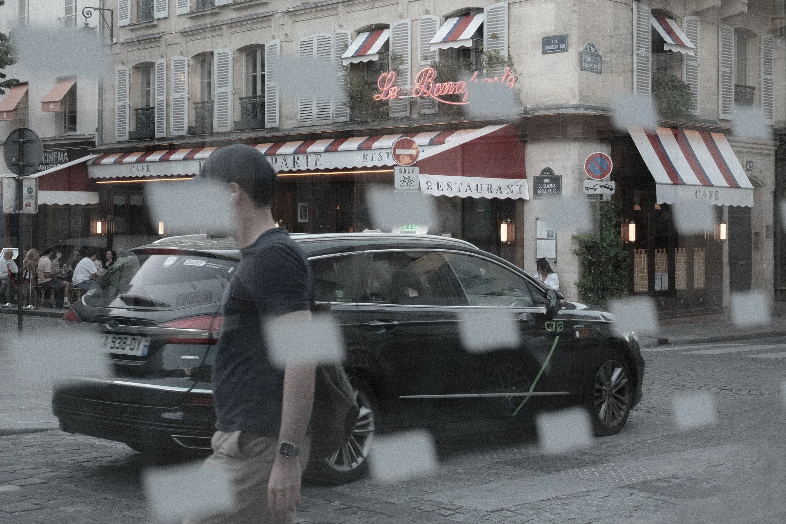 People sitting at outdoor tables of a cafe or restaurant on a city street, with classic European-style buildings in the background, seen through a rain-streaked window.