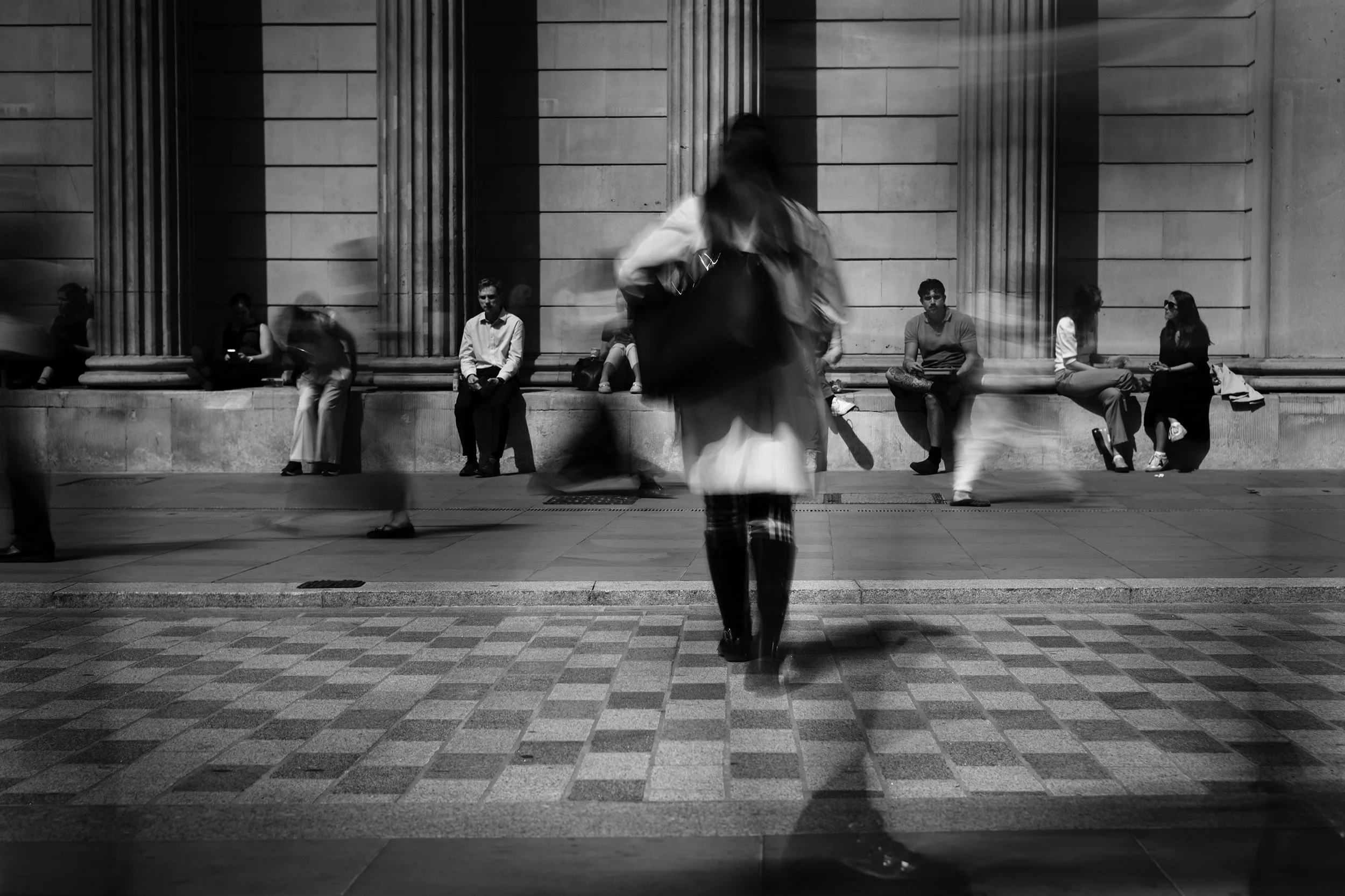 People sitting and standing outside a building with large columns, some blurred walking in front of the camera in black and white.