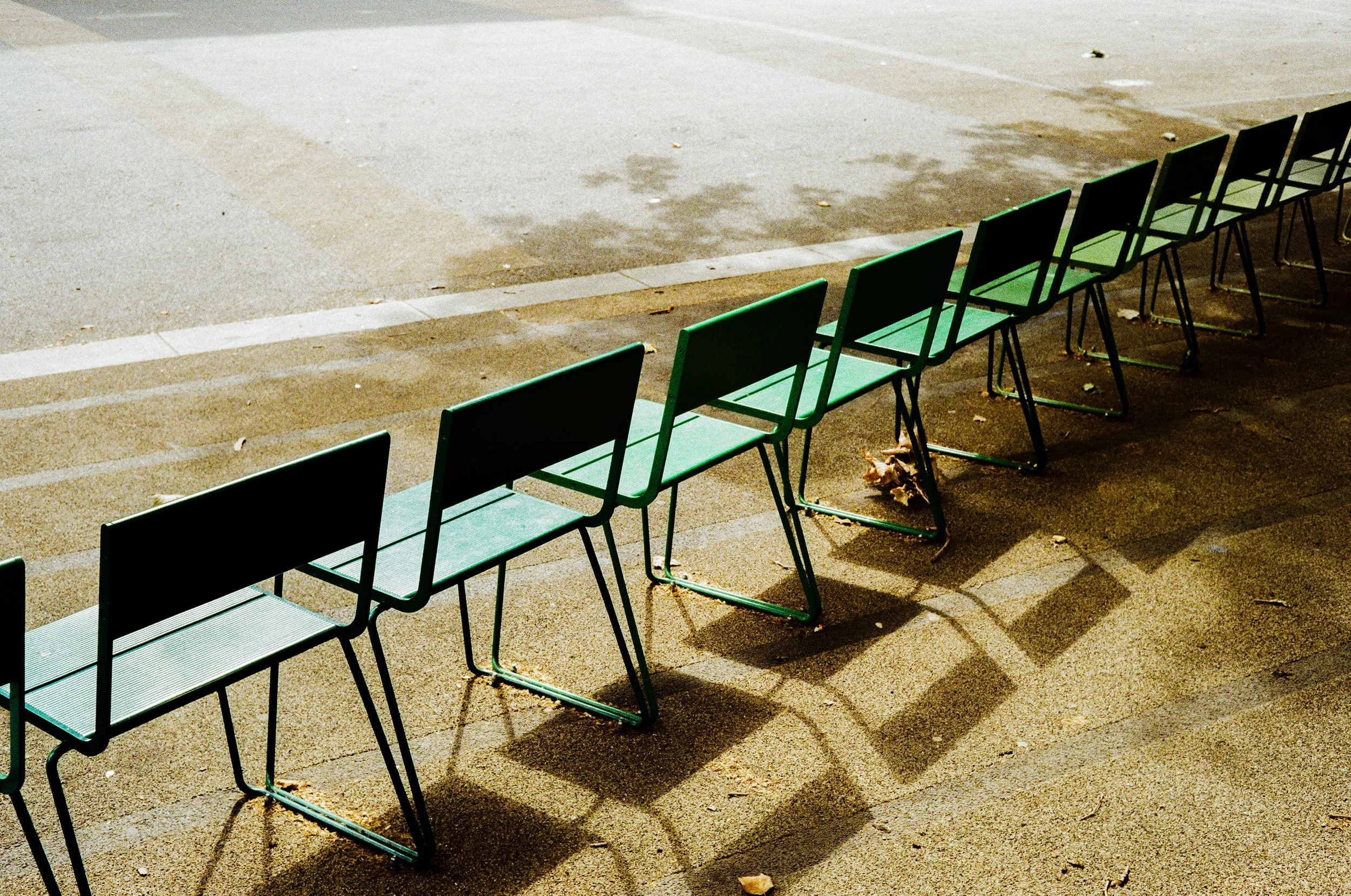A row of green benches lined up in a slight curve, the shadow of a tree is cast upon the asphalt. 