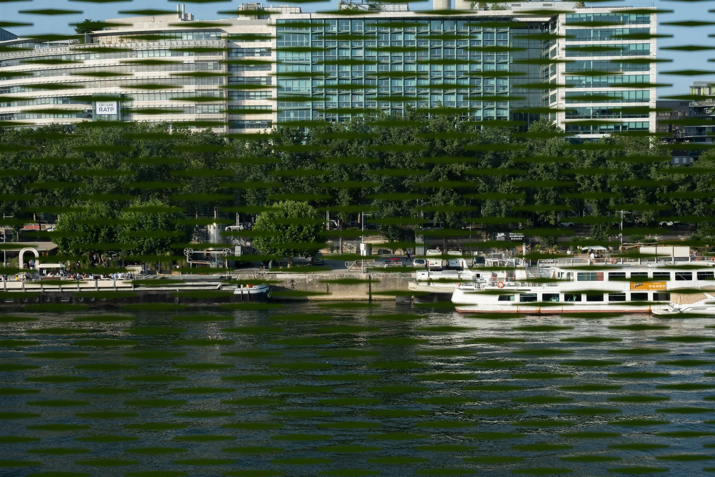 View of a modern multi-story building with numerous windows, partially obscured by green horizontal blinds or reflections, along a waterfront with parked cars, trees, and a boat docked at the shore.
