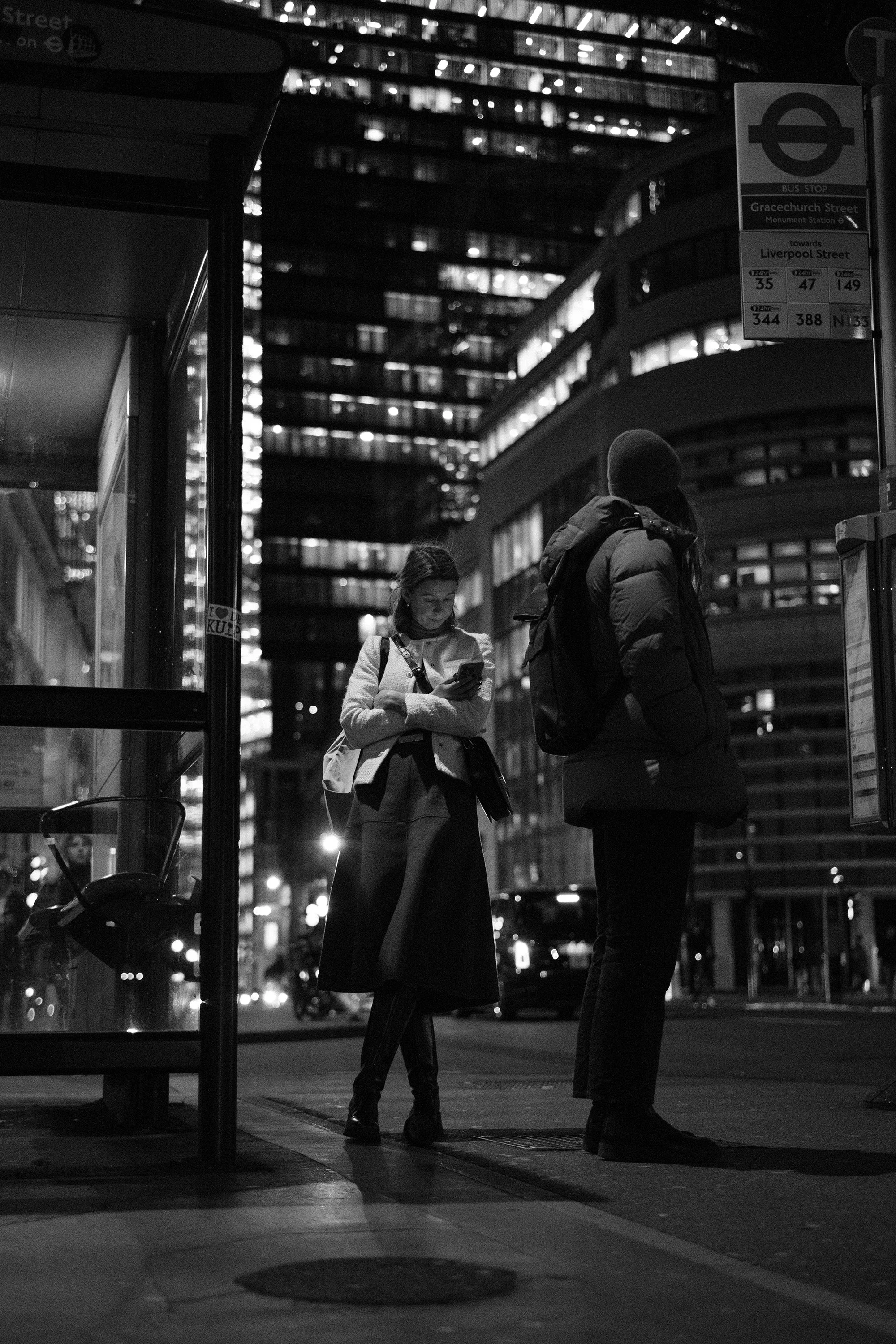Two people standing at a bus stop in an urban area at night, with high-rise buildings in the background. One person is looking at their phone, while the other is facing away. The scene is lit by streetlights and building windows.