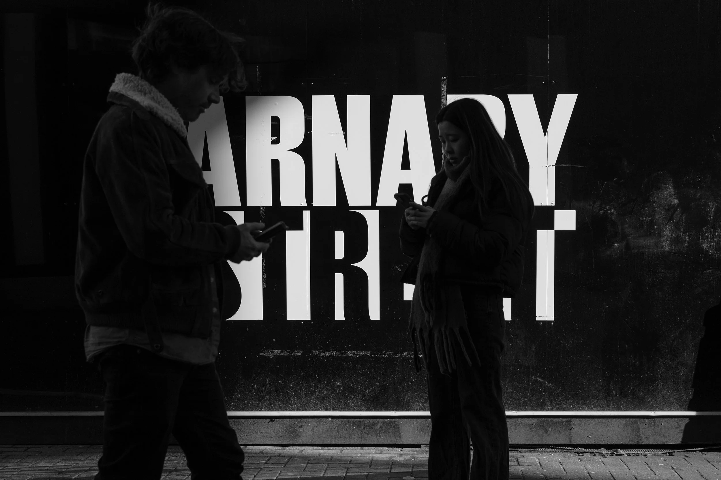 Two people standing in front of a large sign with large text, both looking at their phones.