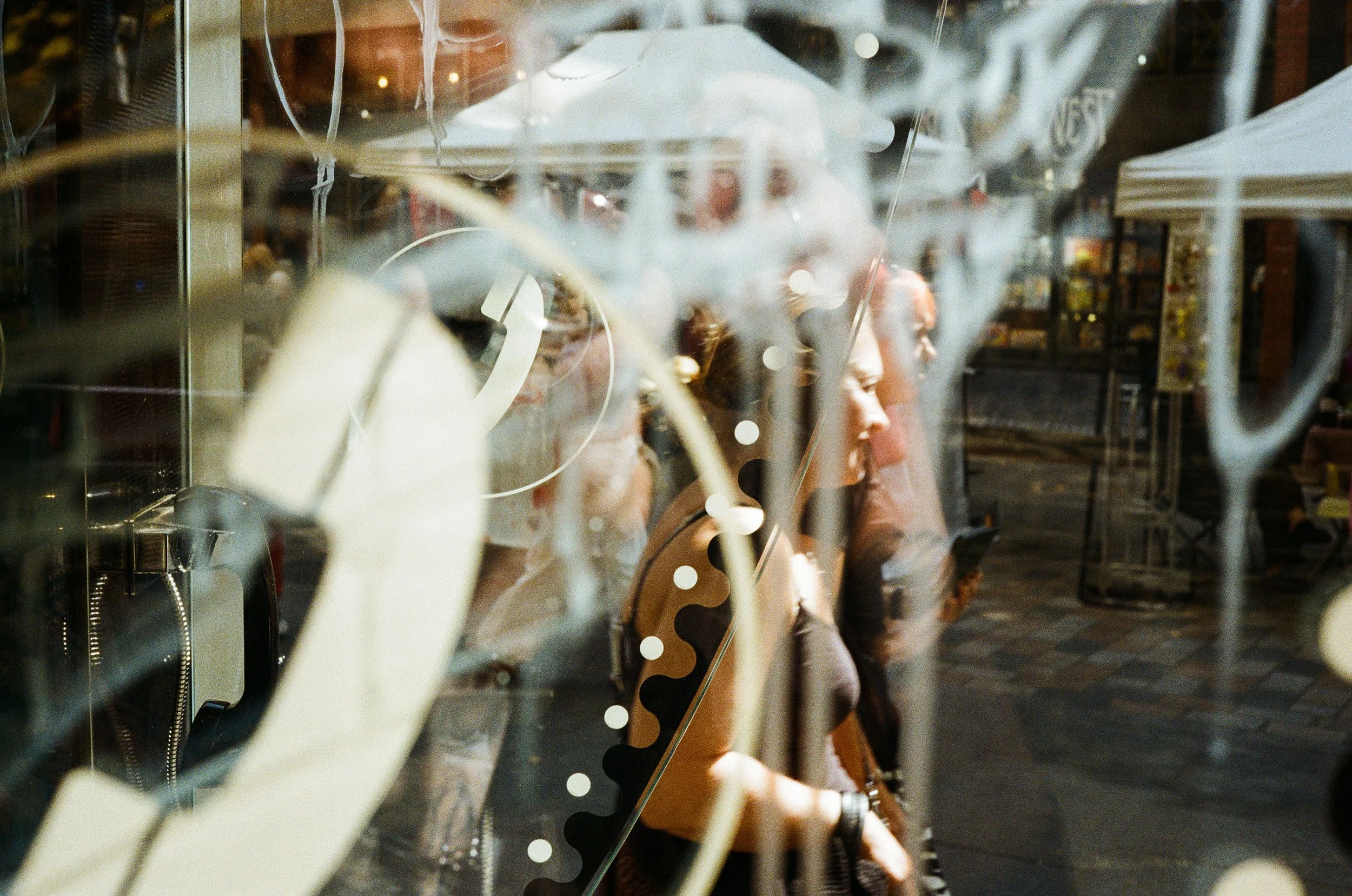 A couple walking behind a public telephone booth with white graffiti written on the booth's windows. The summer sun is illuminating the couple's faces. 