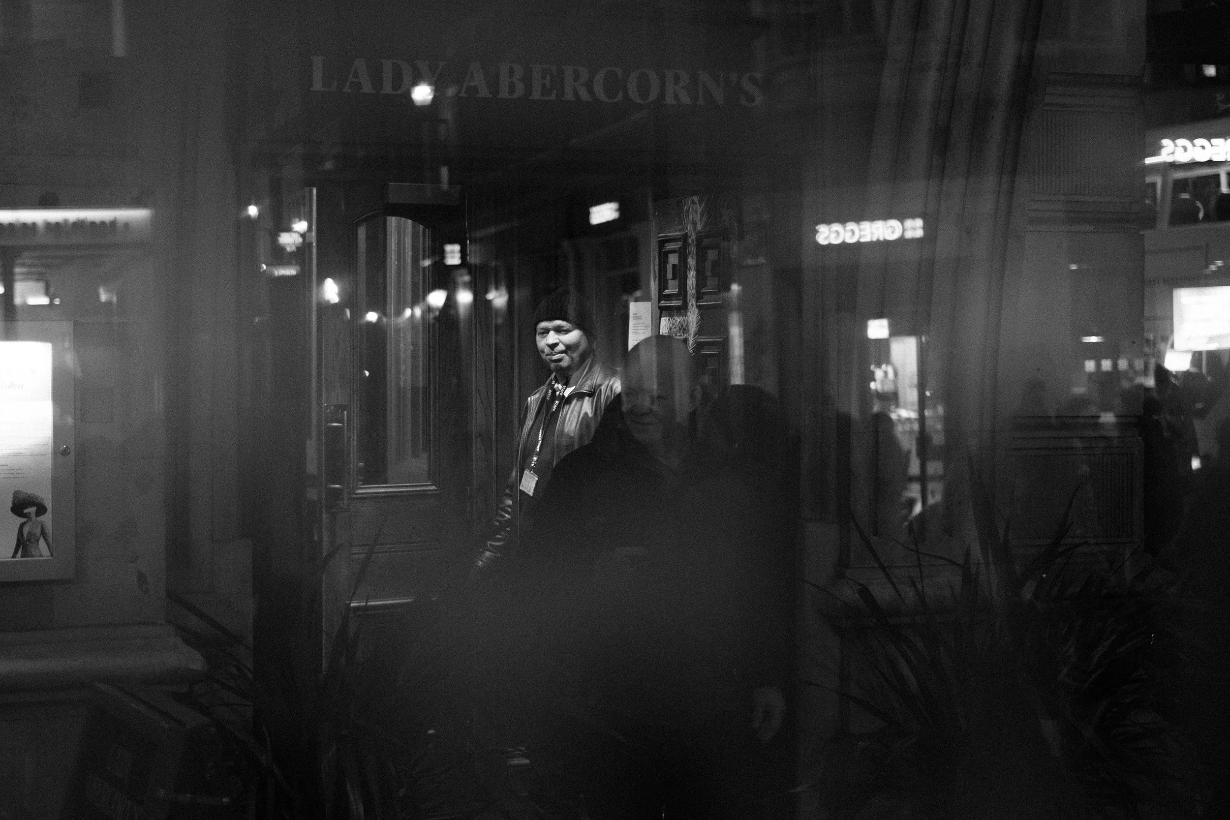 A reflection of two men in a glass window with the word 'Lady Abercorns' above, showing a street scene at night with various illuminated signs.
