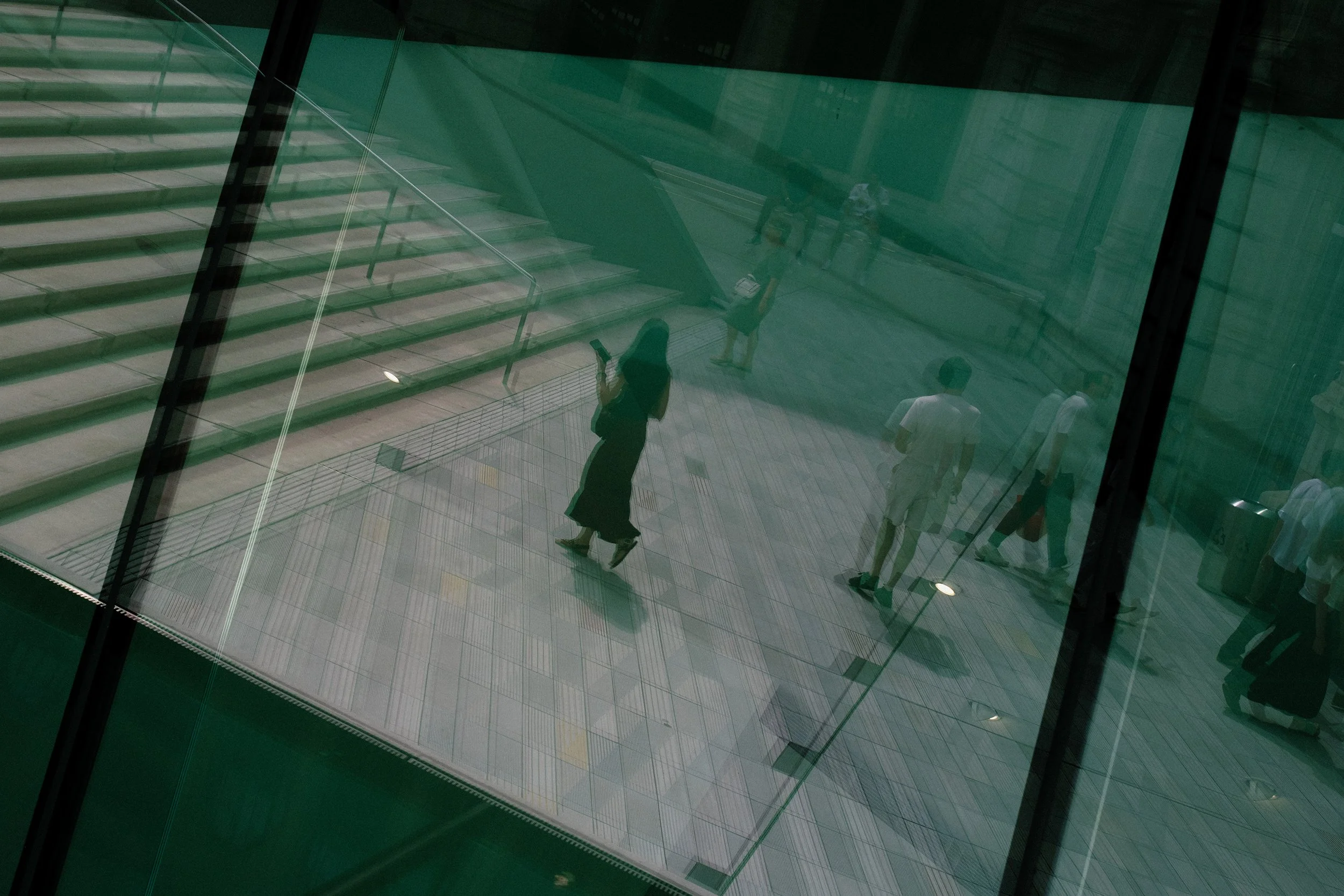View through a glass window of people walking down a staircase and walking across the courtyard, with reflective surfaces and metal railings.