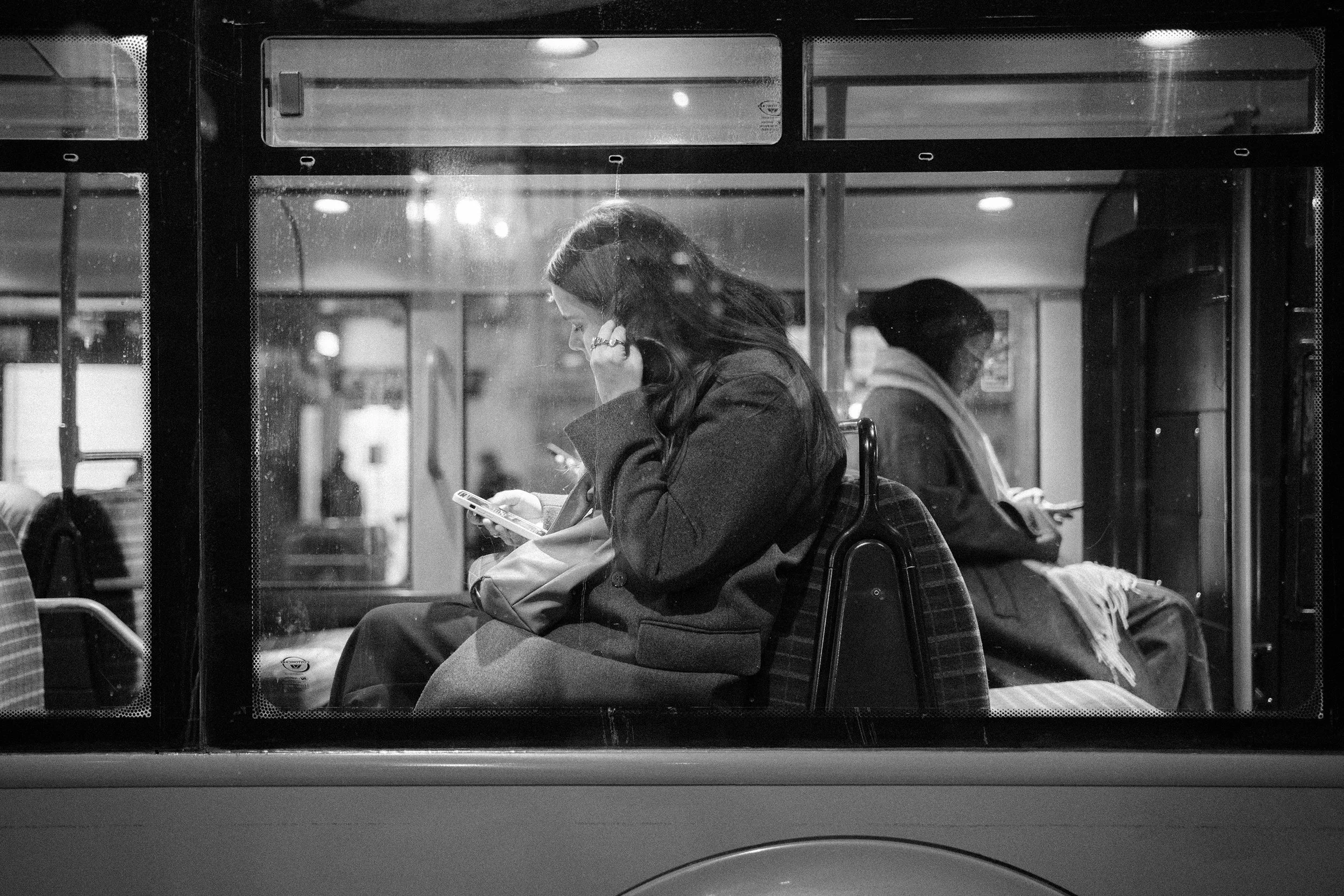 Two women sitting on a bus, both looking at their phones. One is in the foreground with long dark hair, wearing a coat, and holding a bag. The other is in the background, wearing glasses, a scarf, and a coat.