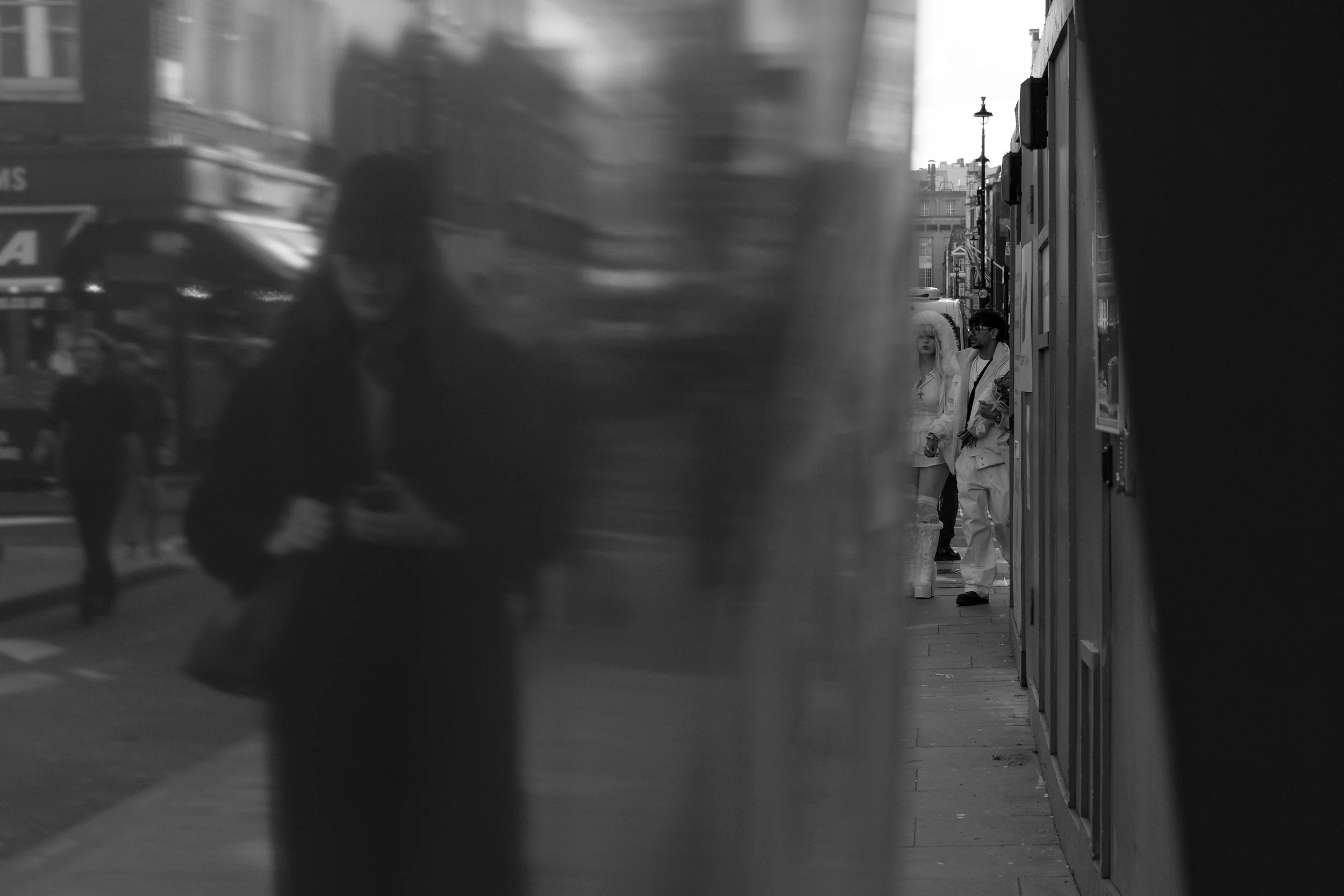 Black and white street scene with two people standing outside a building, one person is wearing a large hooded coat, and the other is holding flowers, with a city background including street lamps and buildings.