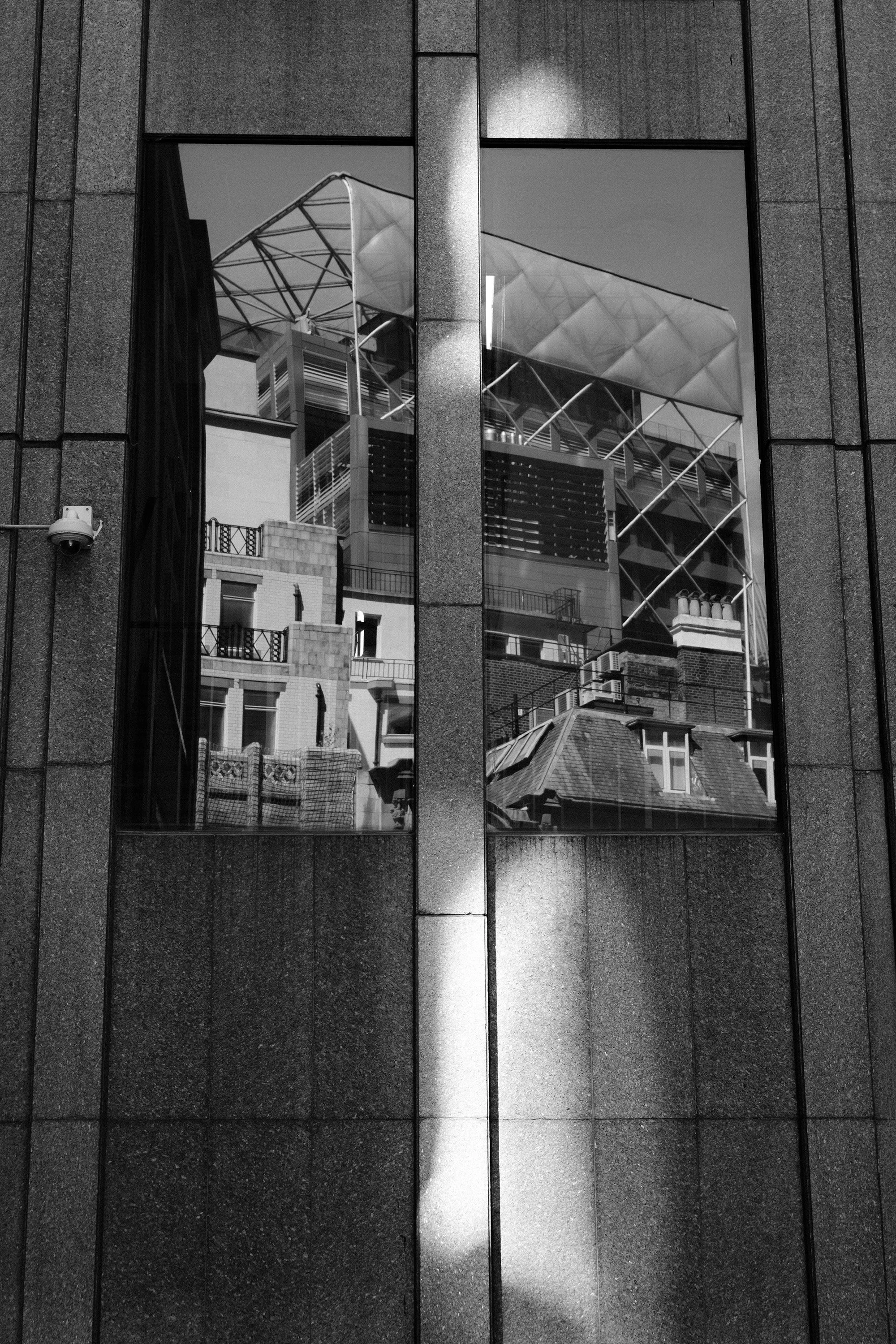 The image shows the reflection of buildings, including a modern glass and steel structure, in a window of a nearby building with a dark stone exterior.