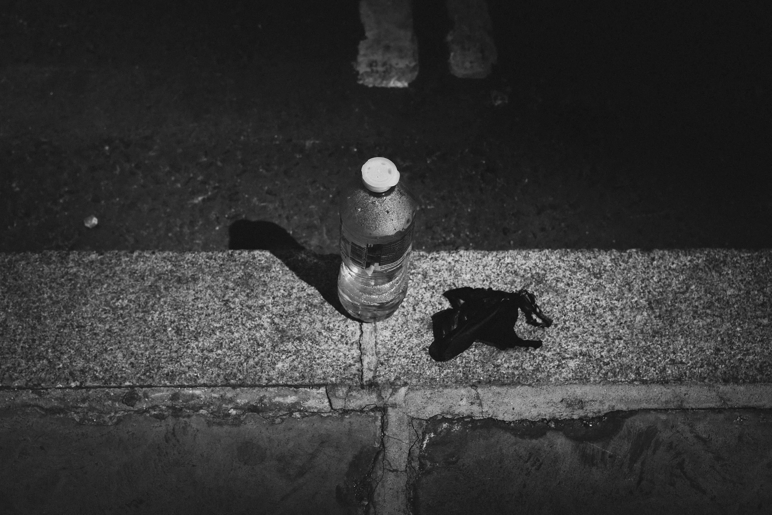 A plastic water bottle with the cap placed on top, positioned on a sidewalk with a shadow cast beneath it. The ground is textured with small gravel and cracks, and there's a dark banana peel on the sidewalk next to the bottle.