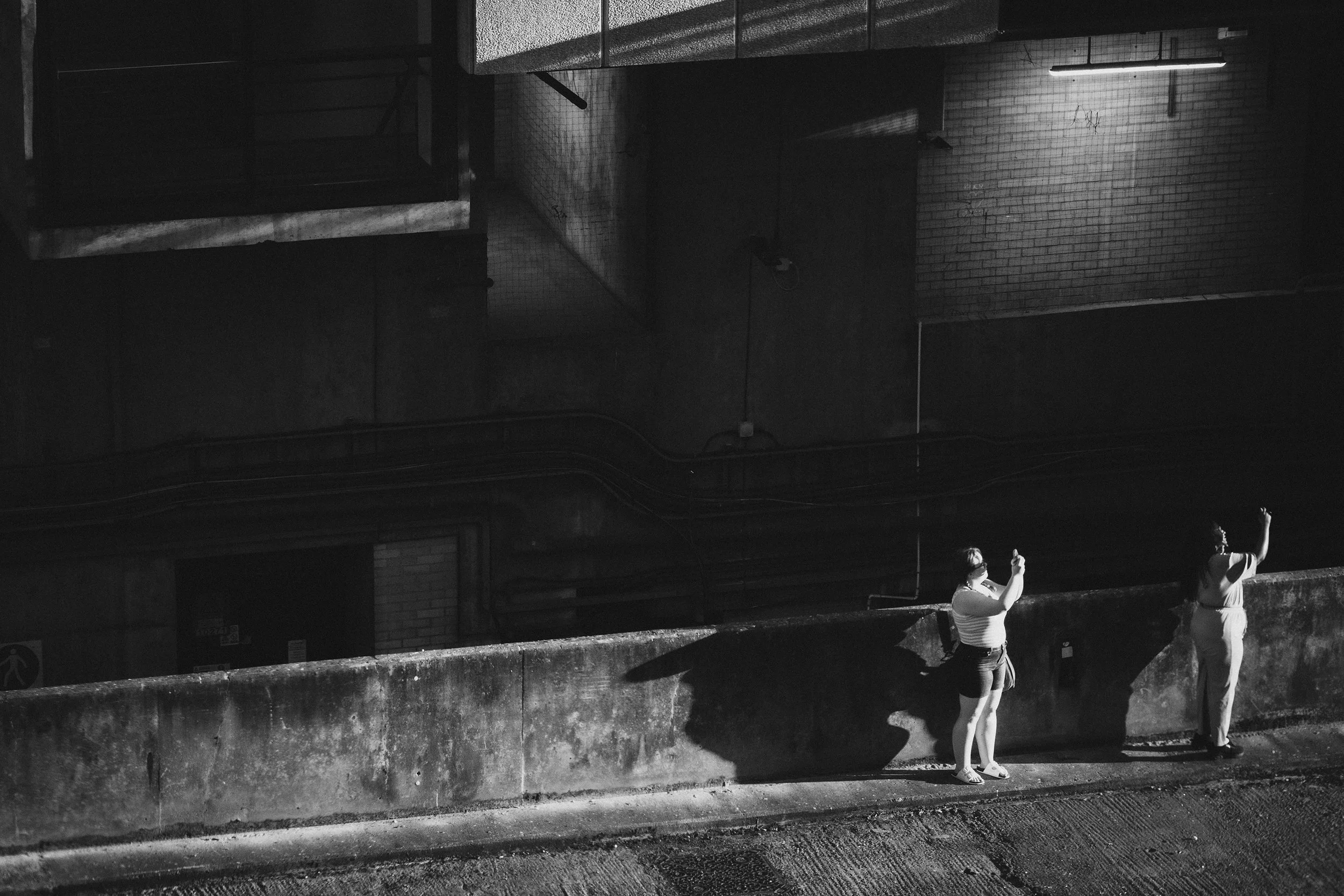 Black and white scene of two women standing on a concrete ledge beside a brick wall, looking up and taking photos with their smartphones.