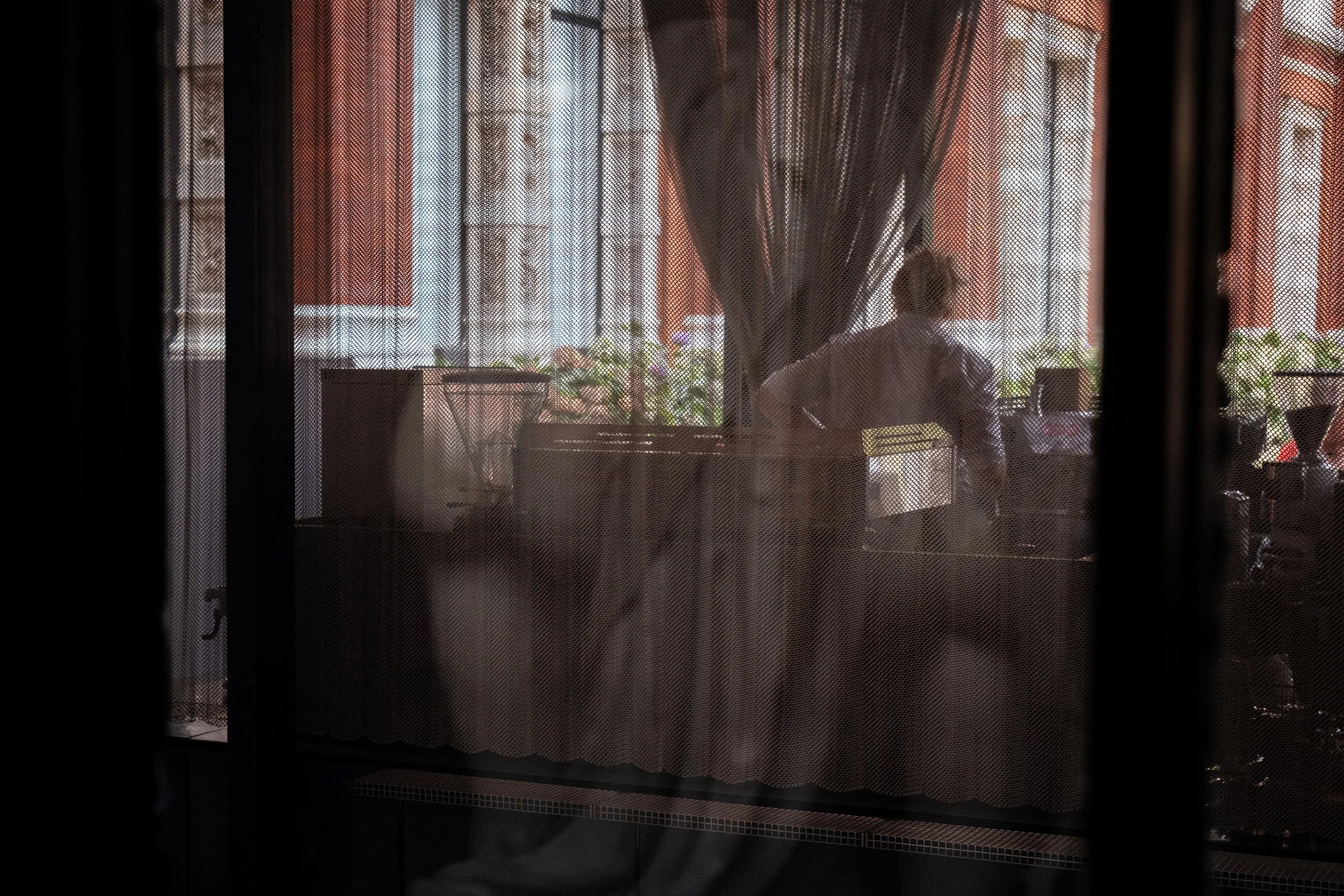 Person standing behind a counter, viewed through a curtain, with buildings and flowers outside the window.