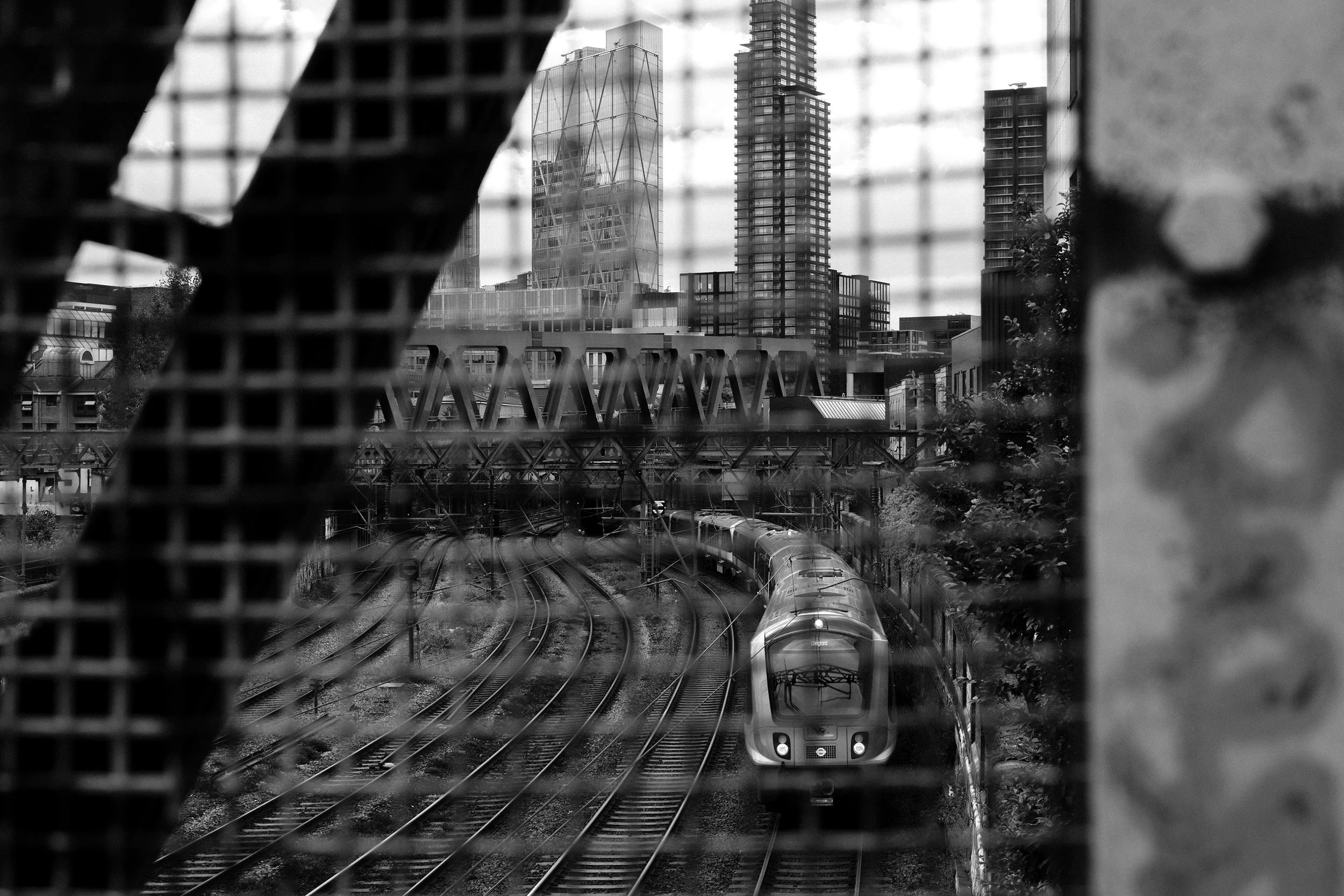 A black and white photo of a train on railway tracks, viewed through a metal grid, with city skyscrapers in the background.