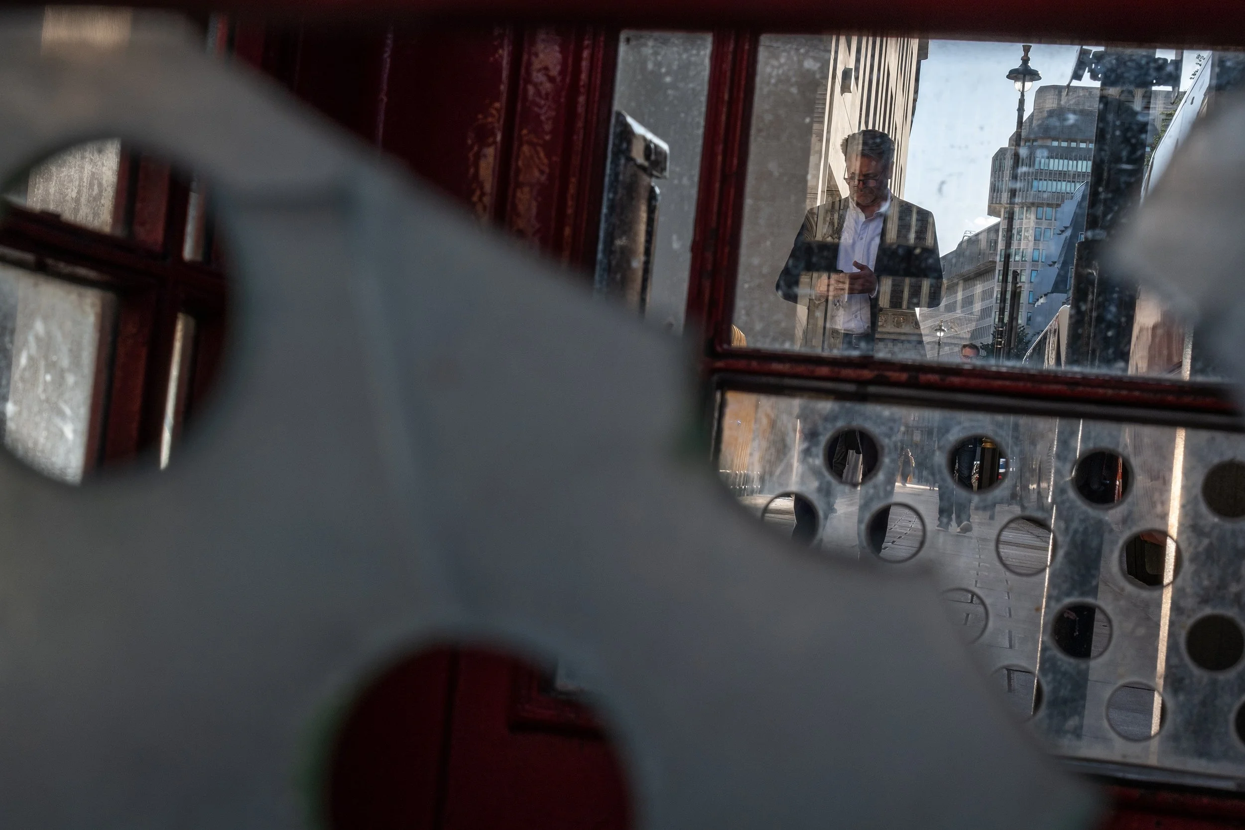 An image shot through a phone booth with broken window panes. A man is looking at his phone, walking towards the phone booth.