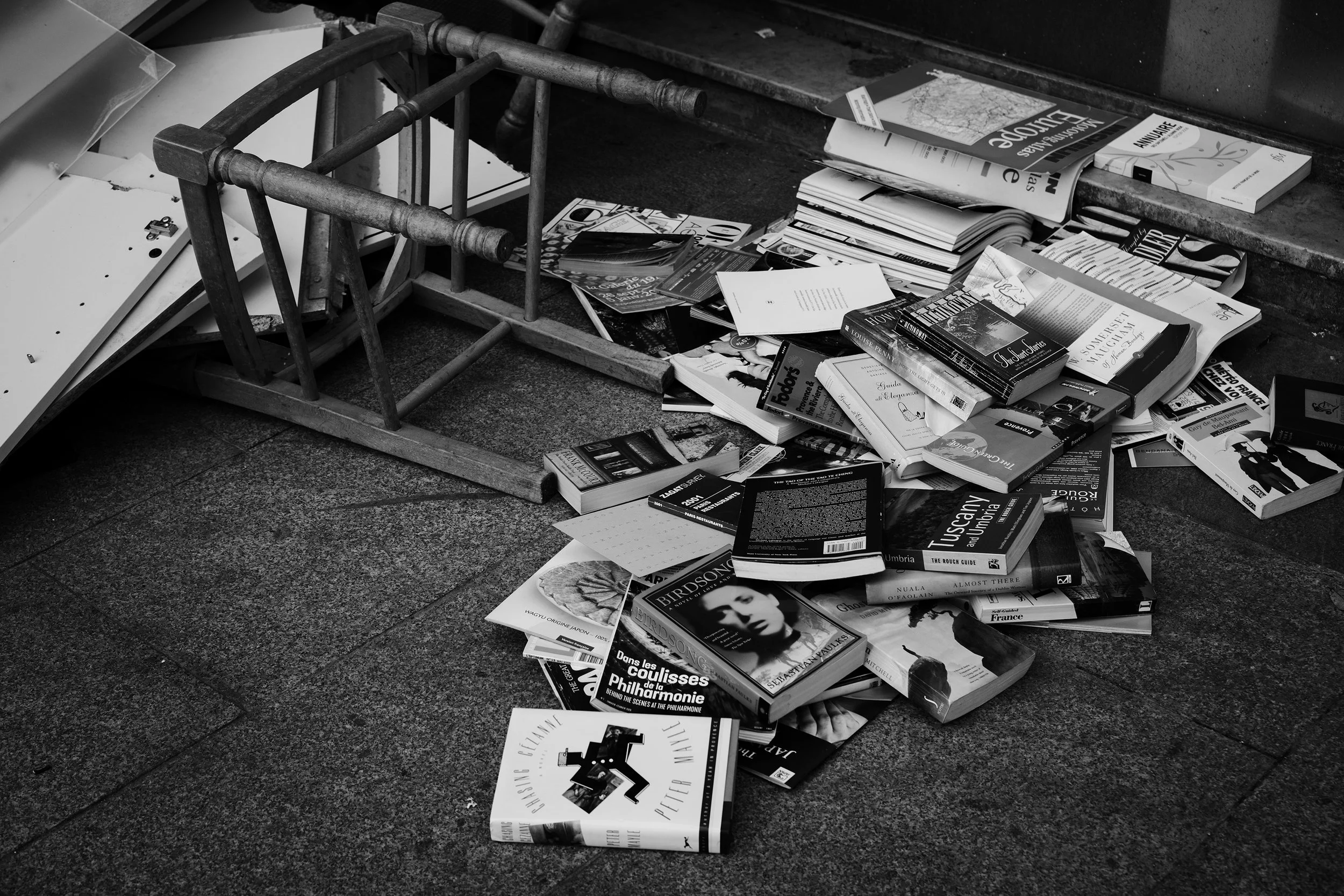 A disorganized pile of magazines and books spilled onto a pavement, with some pages scattered and an overturned wooden chair nearby.