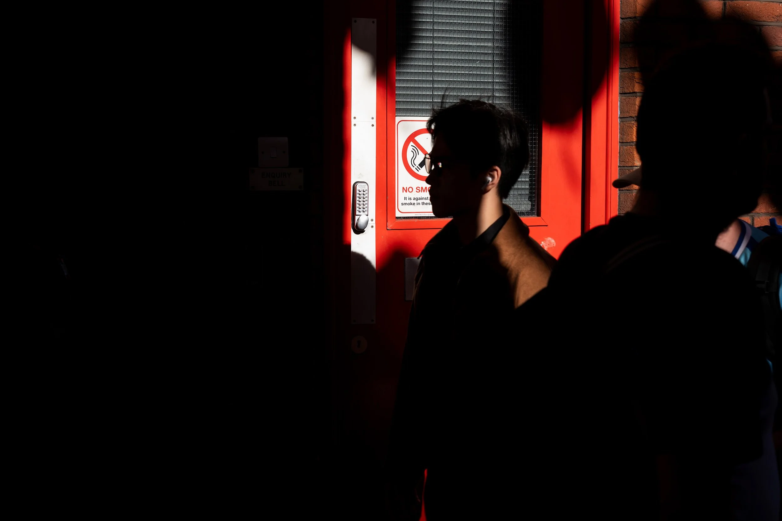 Silhouettes of people walking past a bright red door with a no smoking sign, partly hidden by deep shadows.