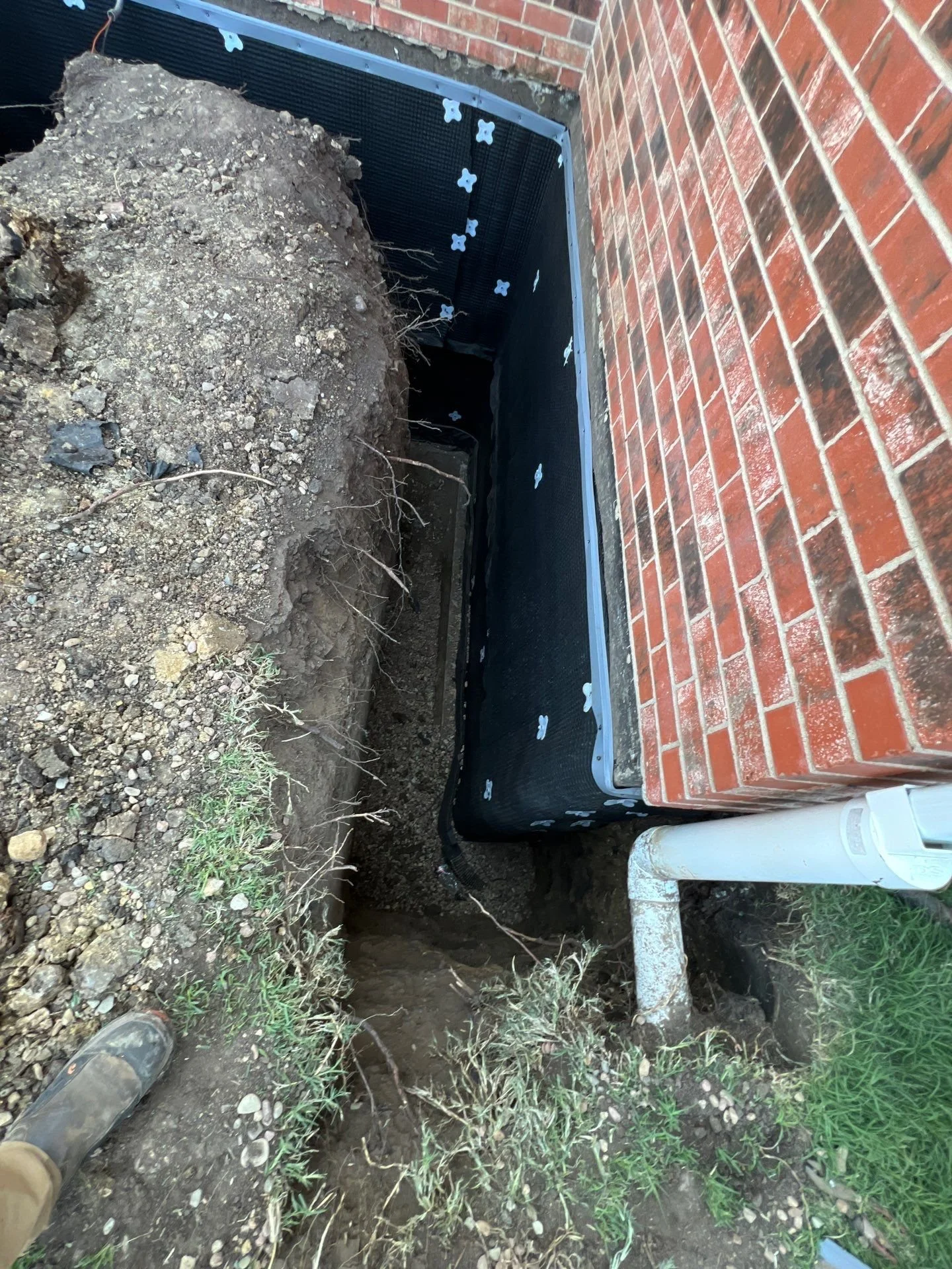 Construction site showing a trench next to a brick house wall with black waterproofing membrane, drain pipe, and soil and grass around the trench.