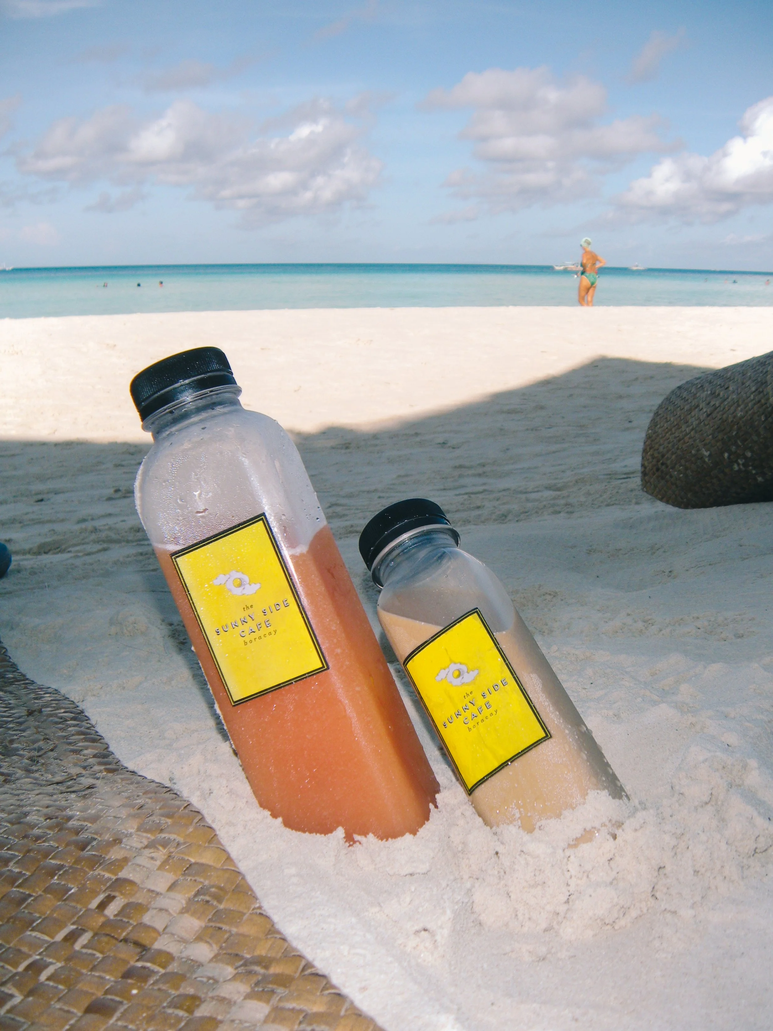 Two plastic bottles filled with fruit beverages resting on sandy beach, with the ocean, cloudy sky, and a person in swimwear in the distance.