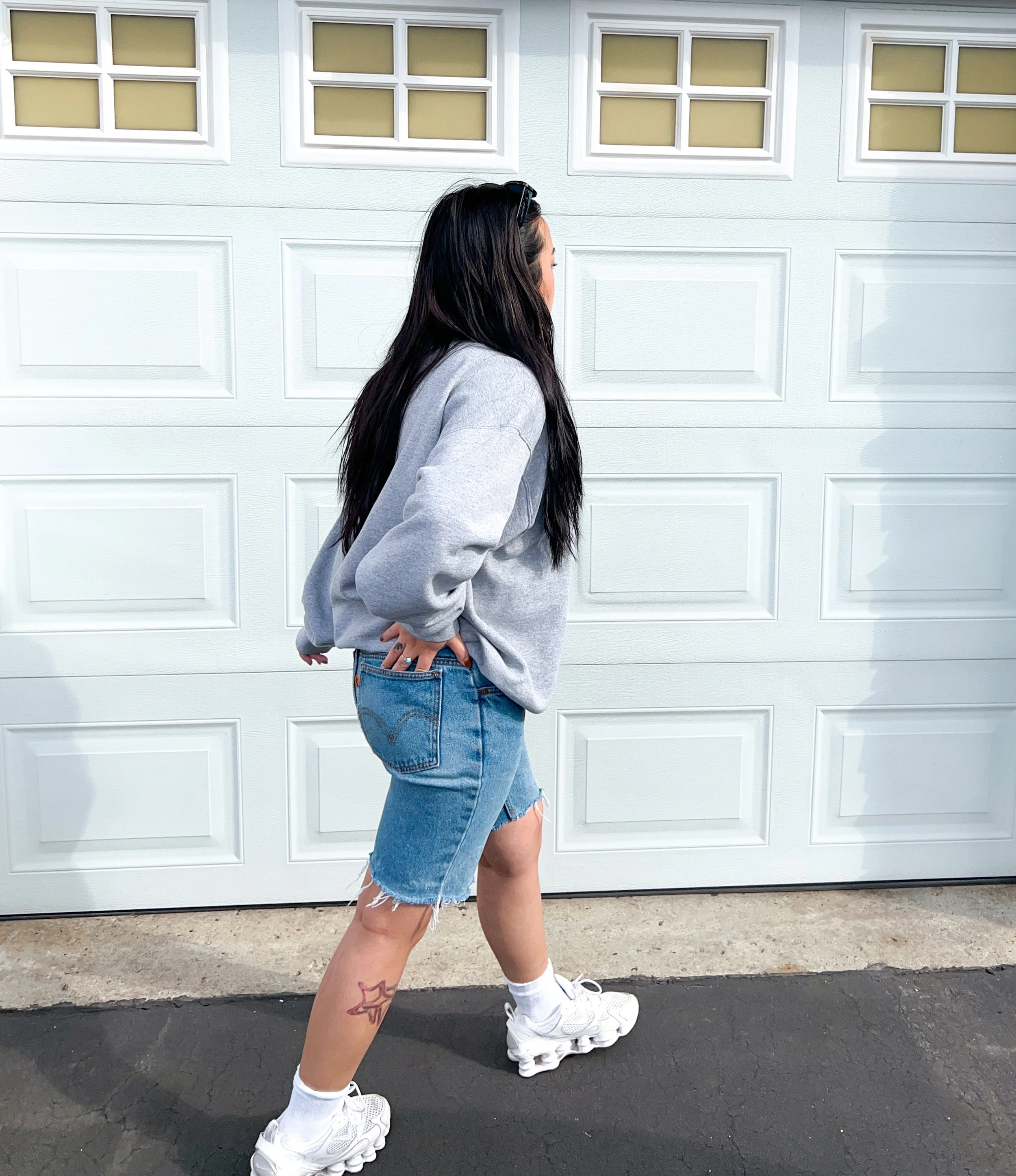 A woman with long dark hair wearing a gray sweatshirt, denim shorts, white sneakers, and white socks standing in front of a white garage door. vintage clothing.