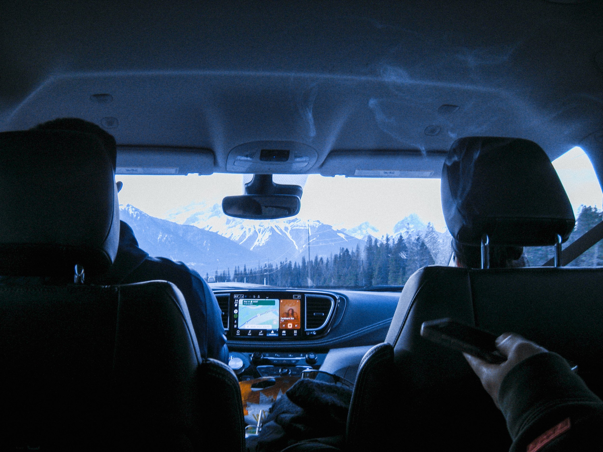 Inside a vehicle with two passengers looking out at snow-capped mountains and a forested landscape through the front windshield.