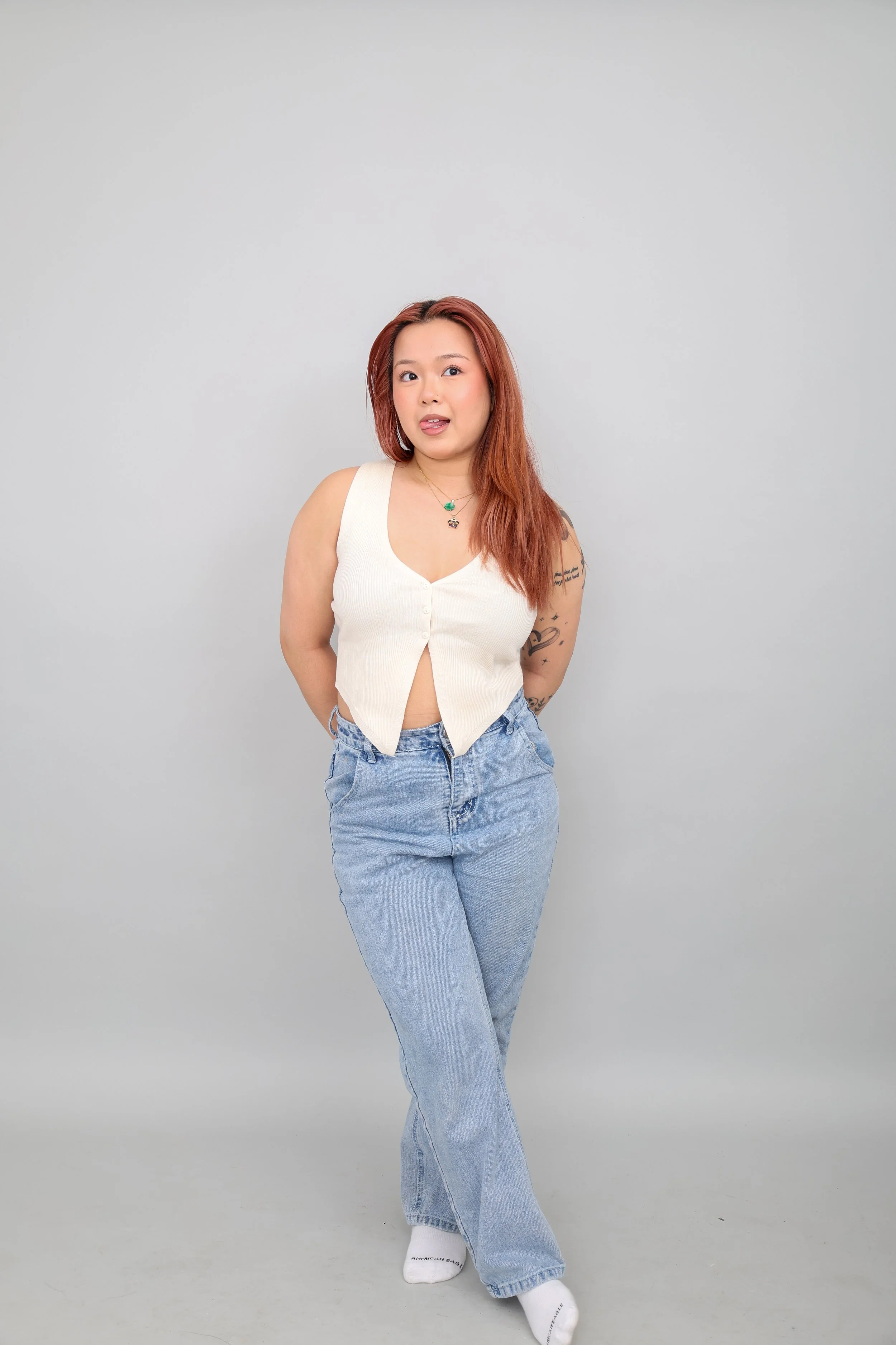 A woman with red hair poses against a plain gray background, wearing a sleeveless white top, high-waisted blue jeans, and white socks. She has tattoos on her right arm and is wearing jewelry, including necklaces.