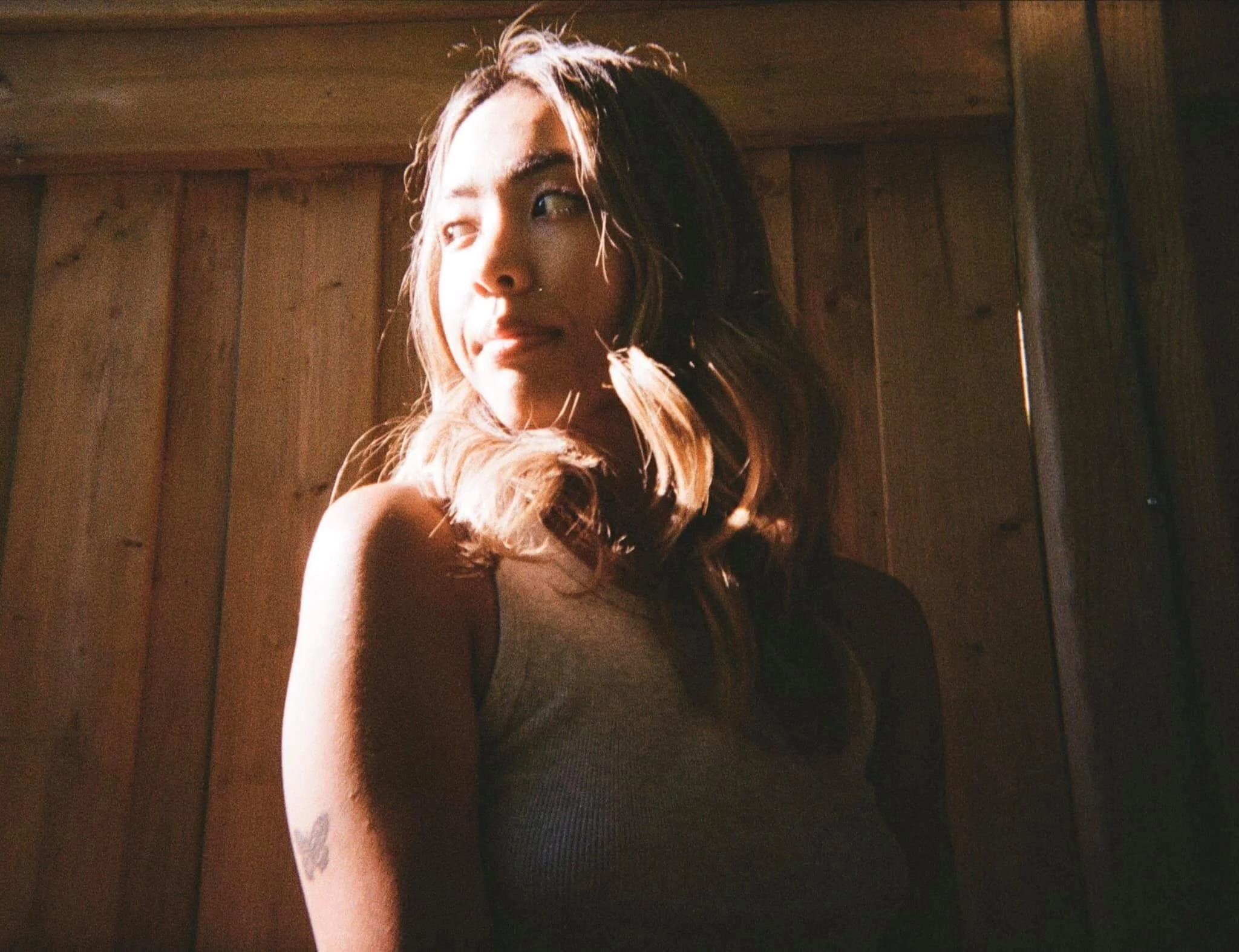 A woman with long wavy hair looking to the side, with sunlight illuminating her face and shoulder, standing in front of a wooden wall.