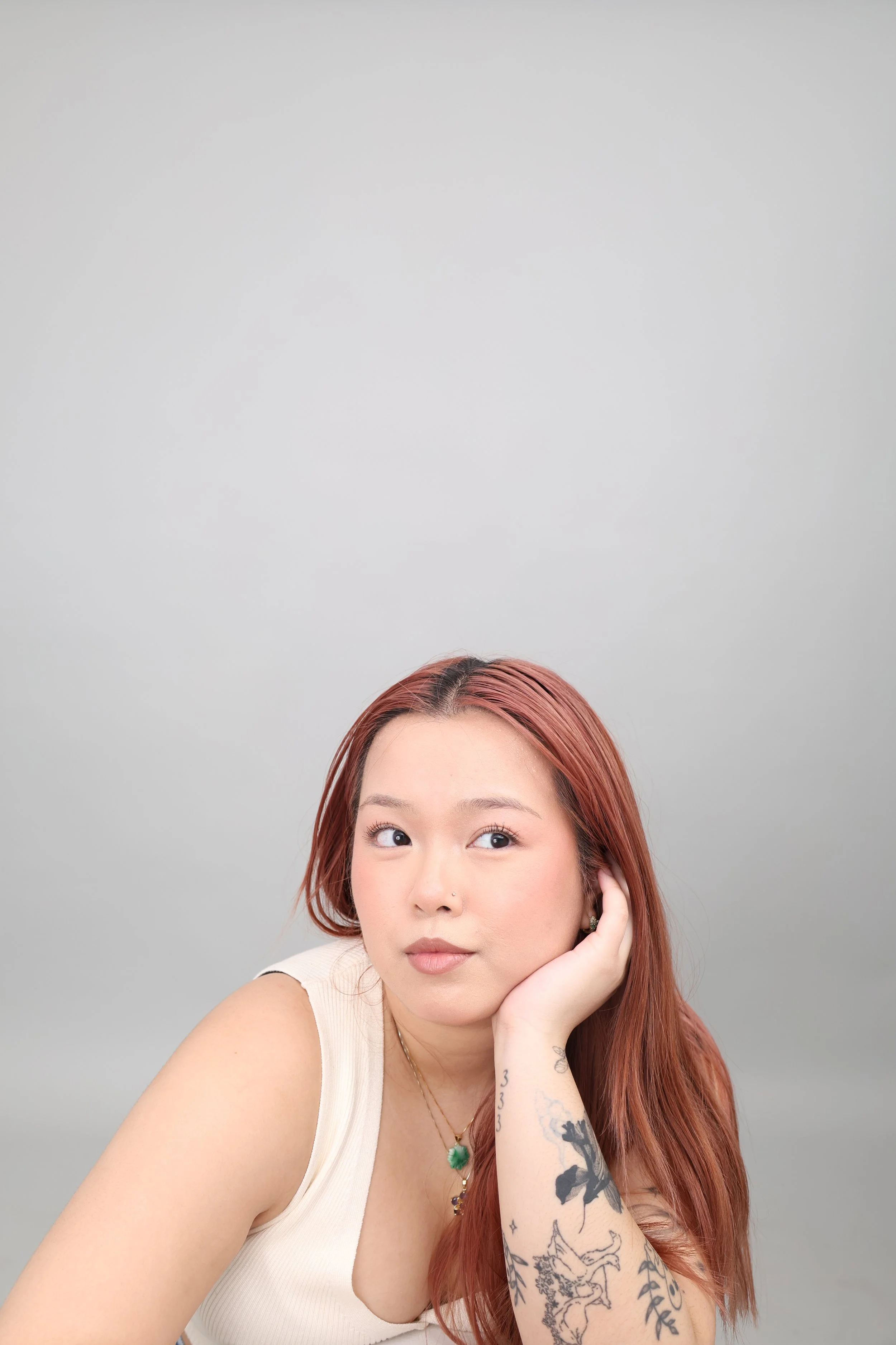A young woman with long reddish hair and tattoos on her arm, resting her chin on her hand while looking slightly to the side, in front of a plain light gray background.