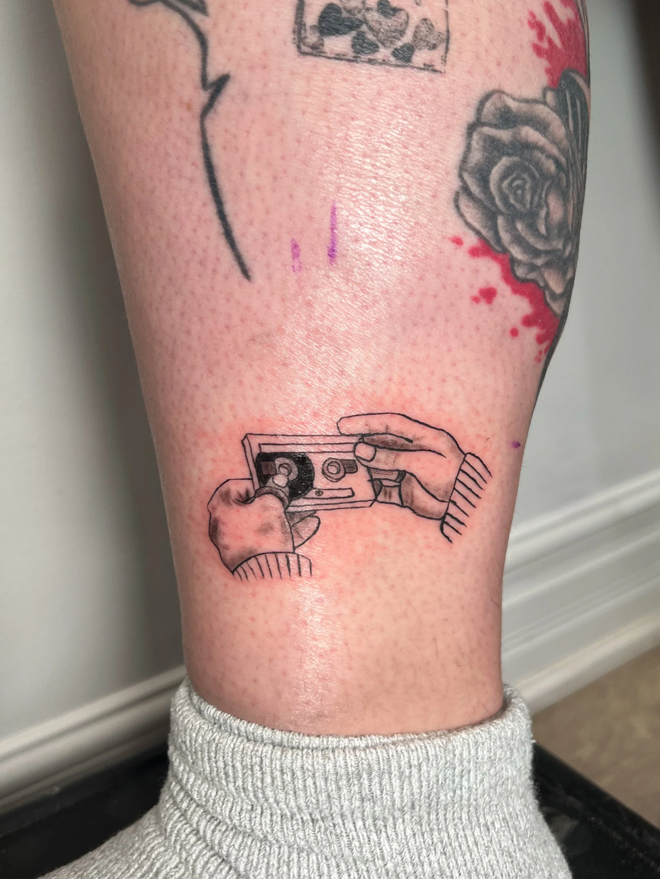 Tattoo of a person holding a cassette tape on the lower leg, with additional tattoos of a rose and other designs visible.