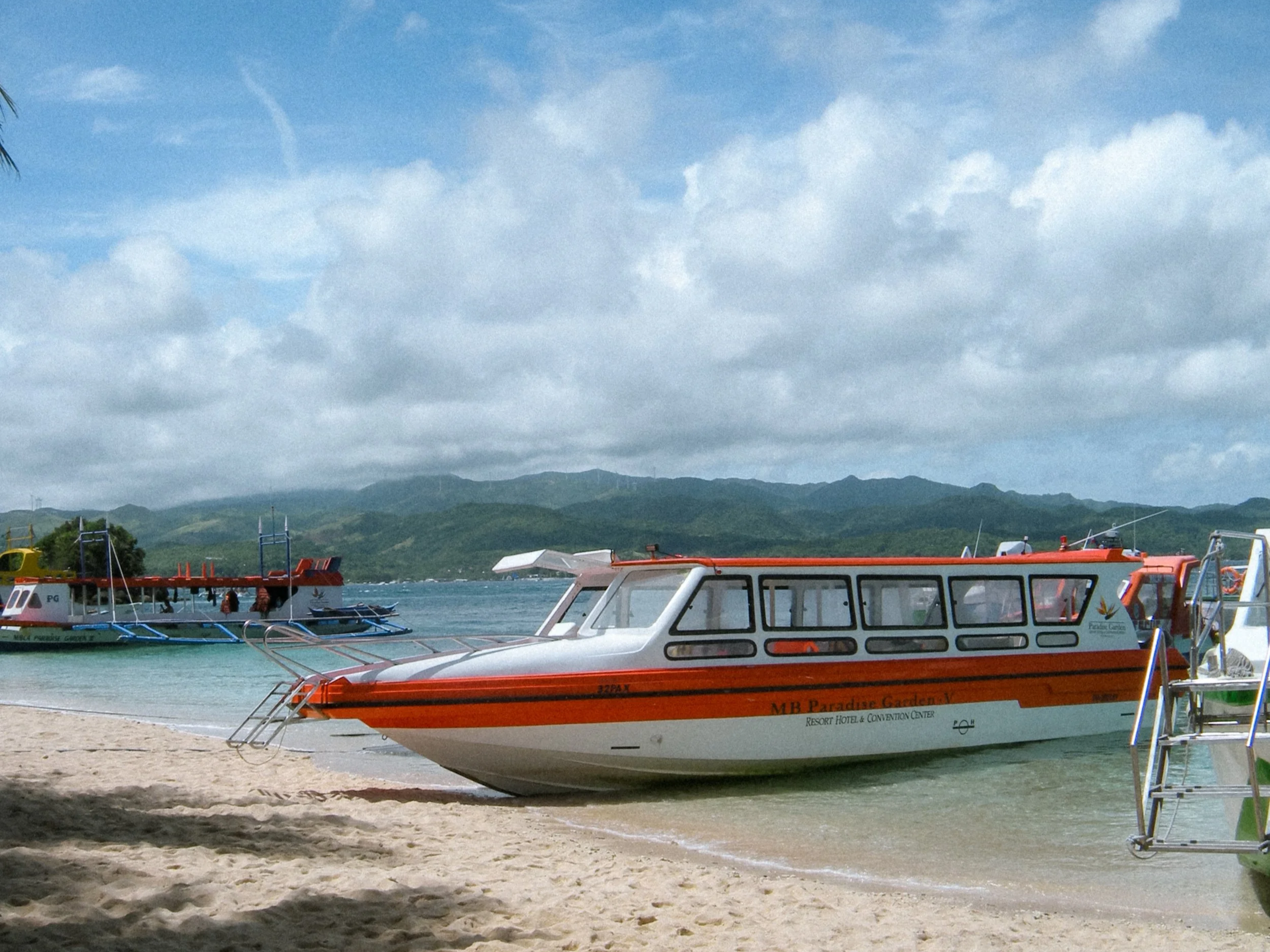 A boat named 'MB Paradise Garden' docked on a sandy beach with other boats in the water, clouds in the sky, and green mountains in the background. boracay..