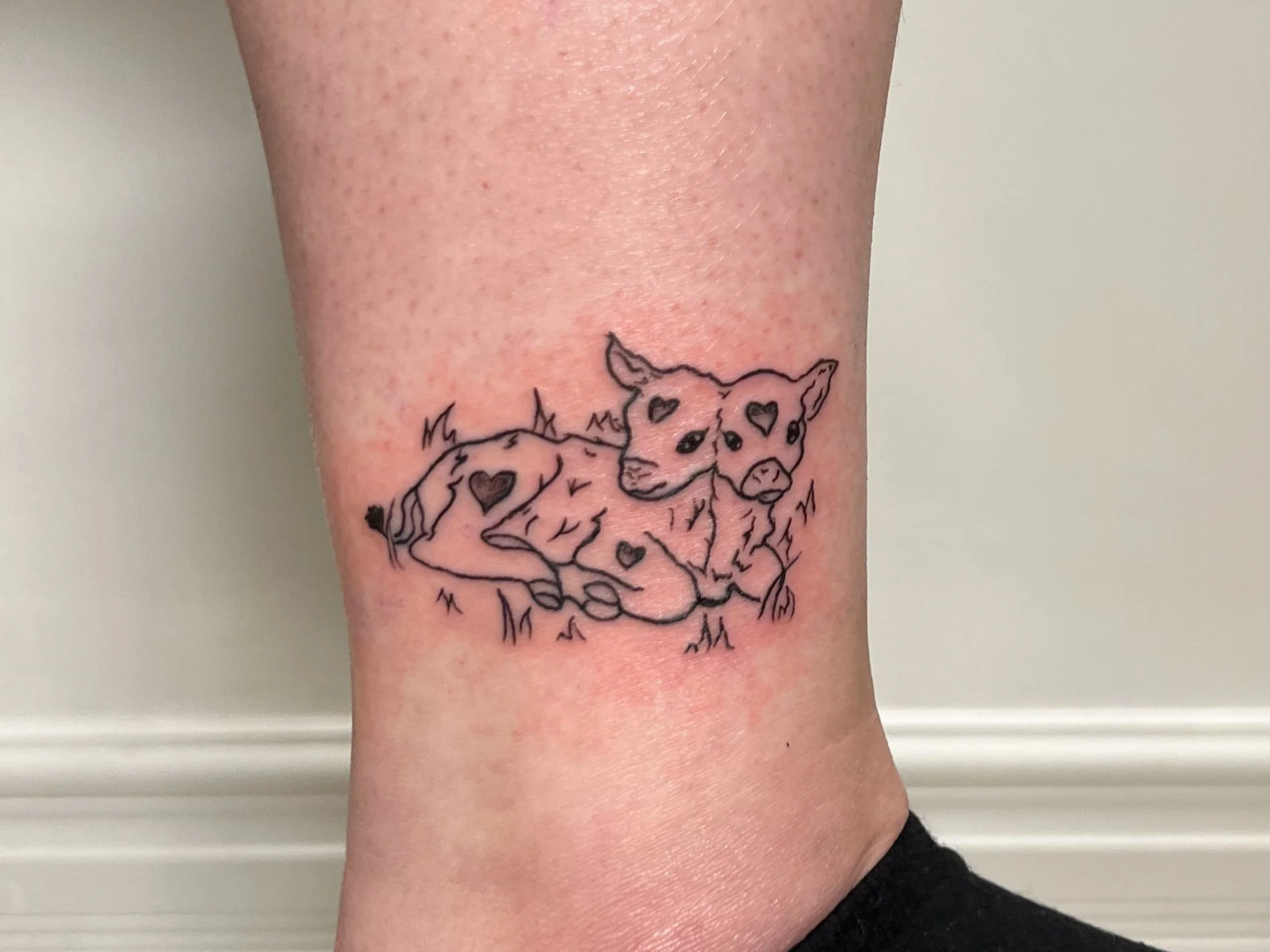 Tattoo of a cute, two-headed calf with heart shapes on its body, lying on grass.