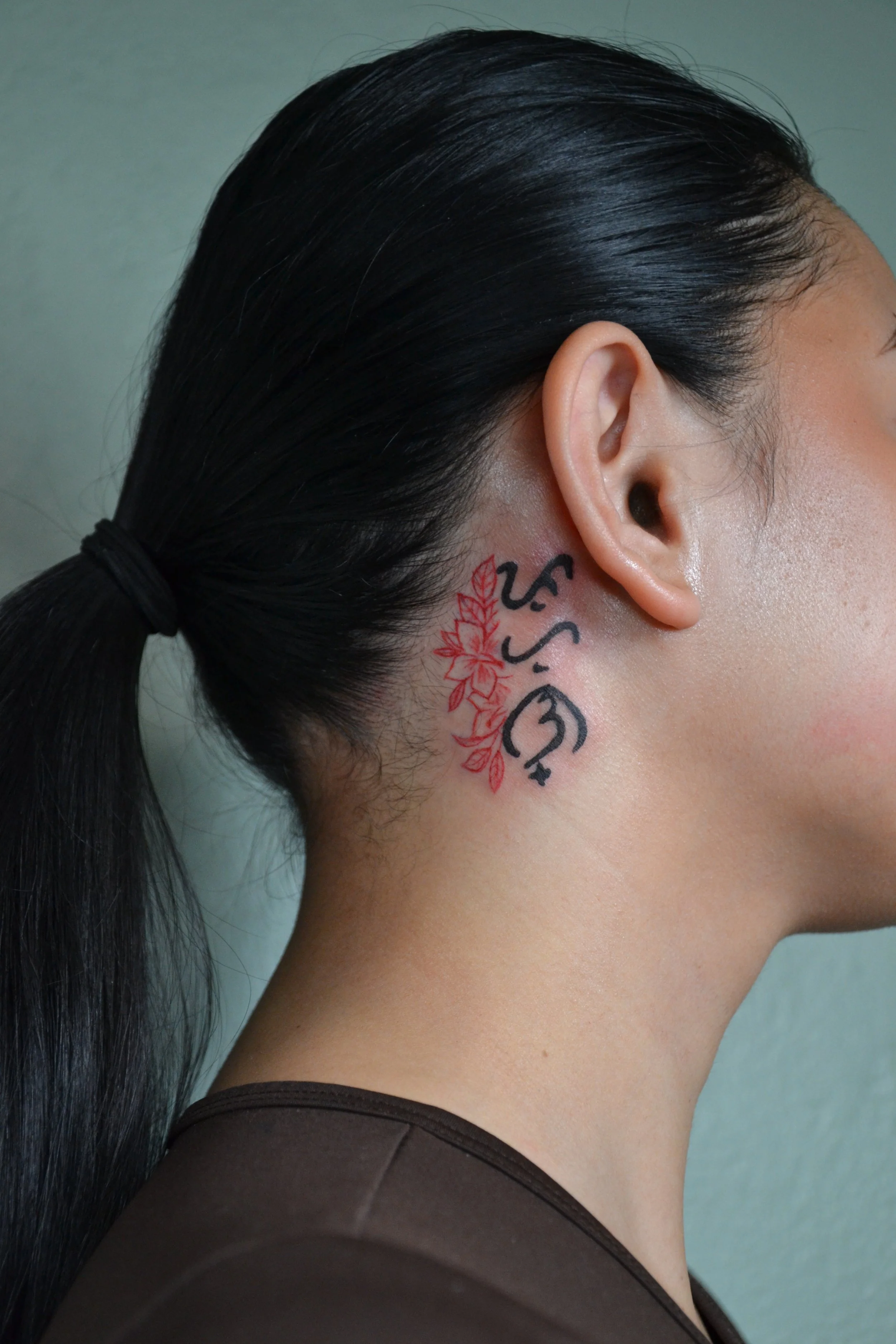 Side profile of a woman with dark hair tied back, showing a tattoo behind her ear with the baybayin text and a floral design in red and black ink.