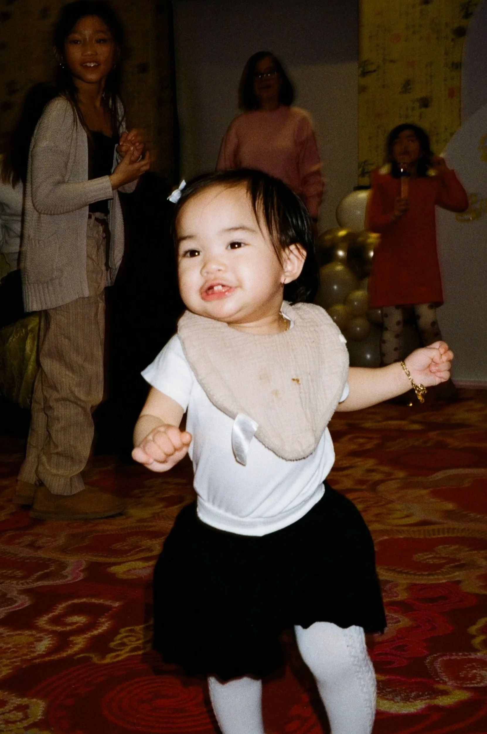 A young girl with dark hair smiling and dancing in the foreground. In the background, three women are standing, with one speaking into a microphone near gold and silver balloons, at an indoor event with a patterned carpet.