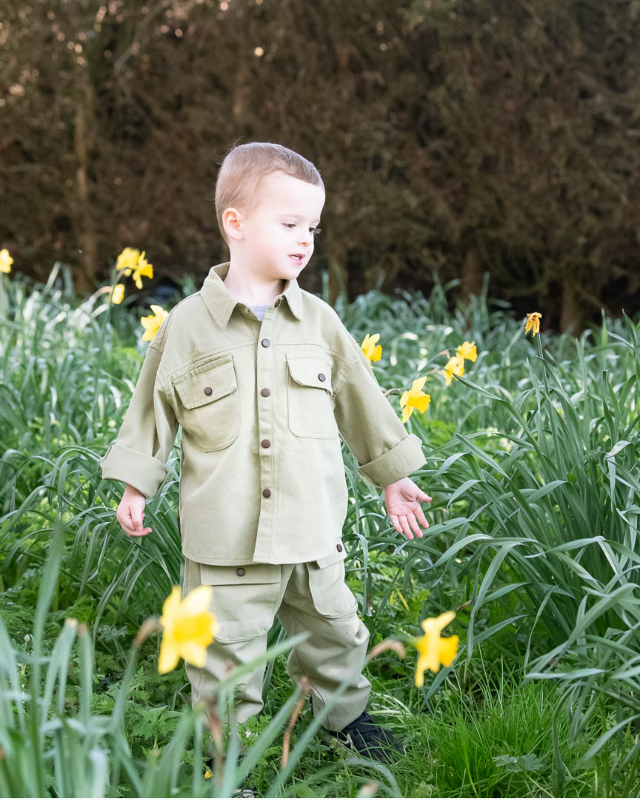 A young boy in beige jacket and pants standing among yellow flowers in a garden with green foliage and trees in the background.