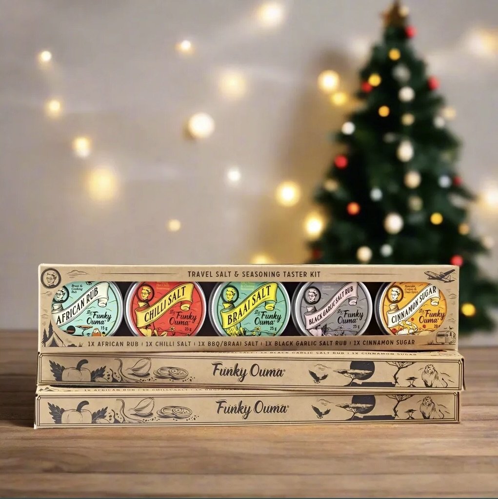 A travel salt and seasoning taster kit with five labeled tins, including African Rub, Chilli Salt, Brava Salt, Black Garlic Salt Rub, and Cinnamon Sugar, stacked on a wooden surface with a decorated Christmas tree in the background.