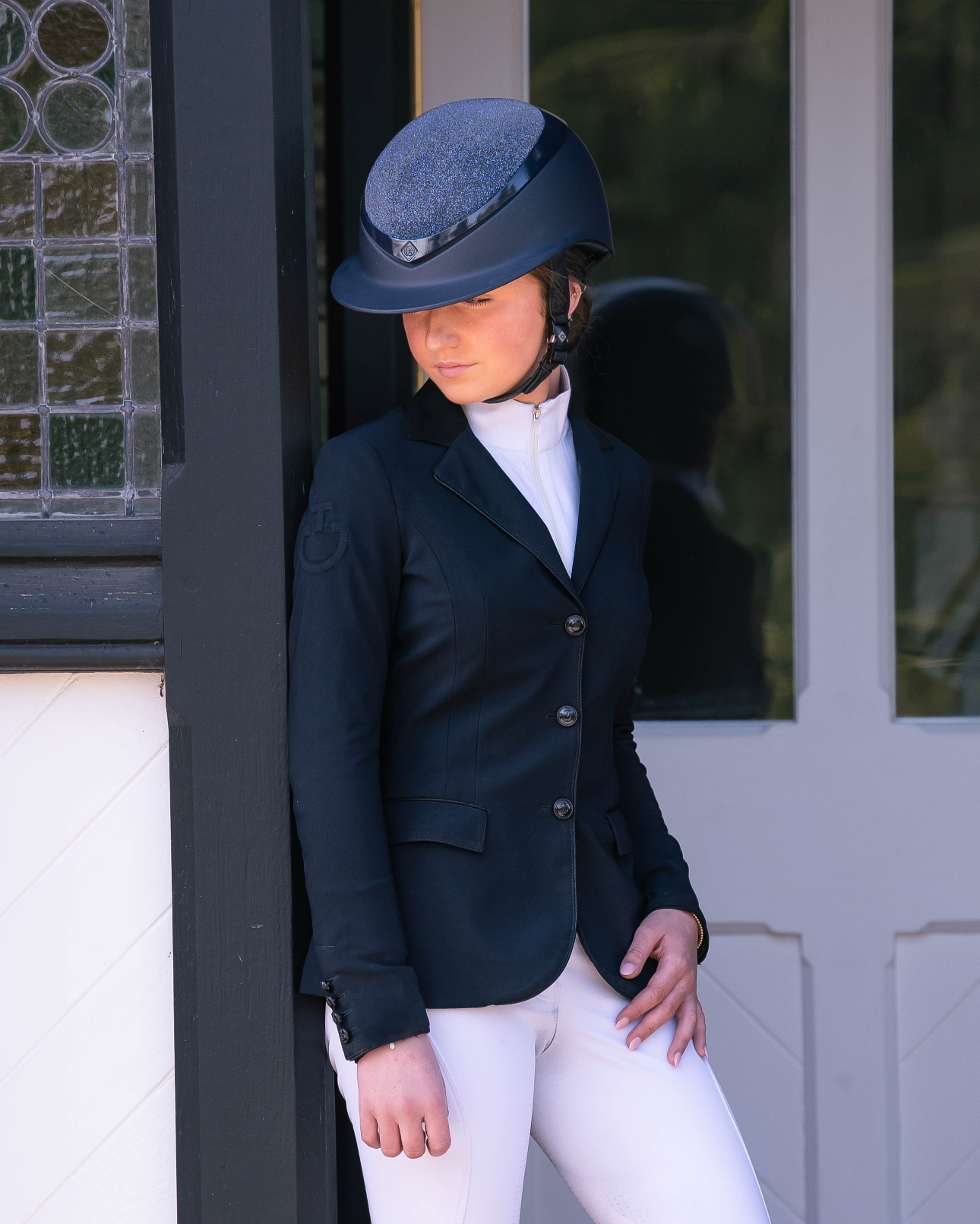 A woman wearing a black equestrian helmet, a black riding jacket, a white shirt, and white riding pants, leaning against a black and white wall outside a building with a glass window and door.