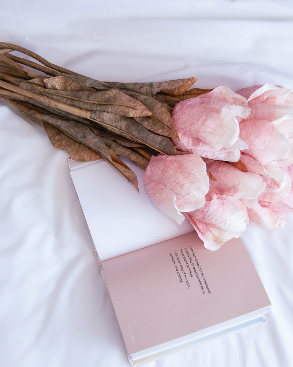 A bouquet of pink and white artificial flowers with brown leaves resting on an open book with a handwritten note, all on a white surface.